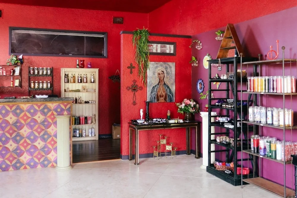Interior of a colorful spiritual shop with red and purple painted walls, shelves with various candles and supplies, a framed picture of the Virgin Mary, and a counter with patterned tile.