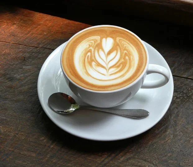 A cup of latte with latte art on top, sitting on a white saucer with a small spoon, on a wooden table.