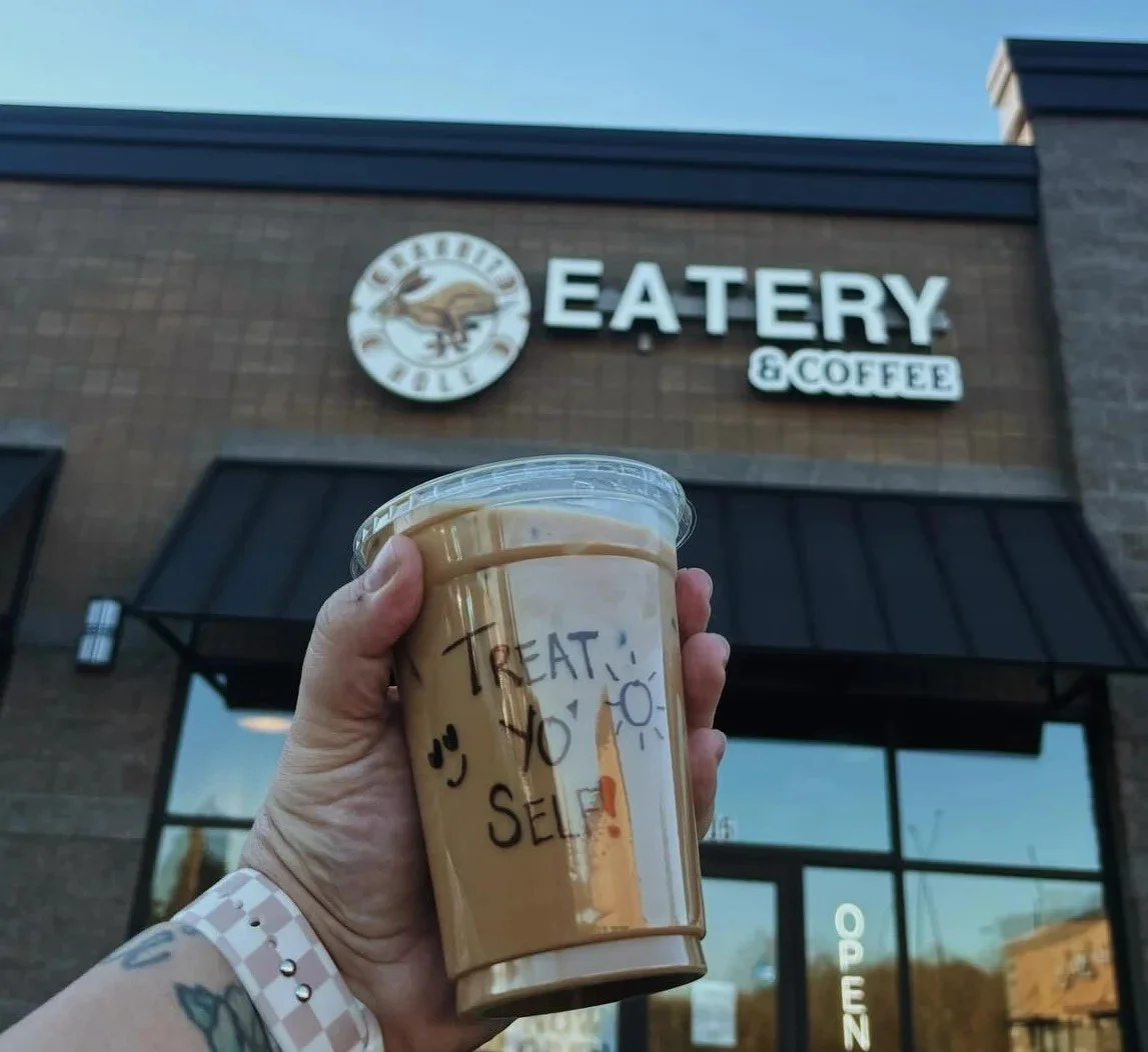 Hand holding a plastic cup of coffee with handwritten message 'TREAT YO SELF' outside a restaurant called 'GEOFFREY'S EATER' with logo, brick building, and a glass door.