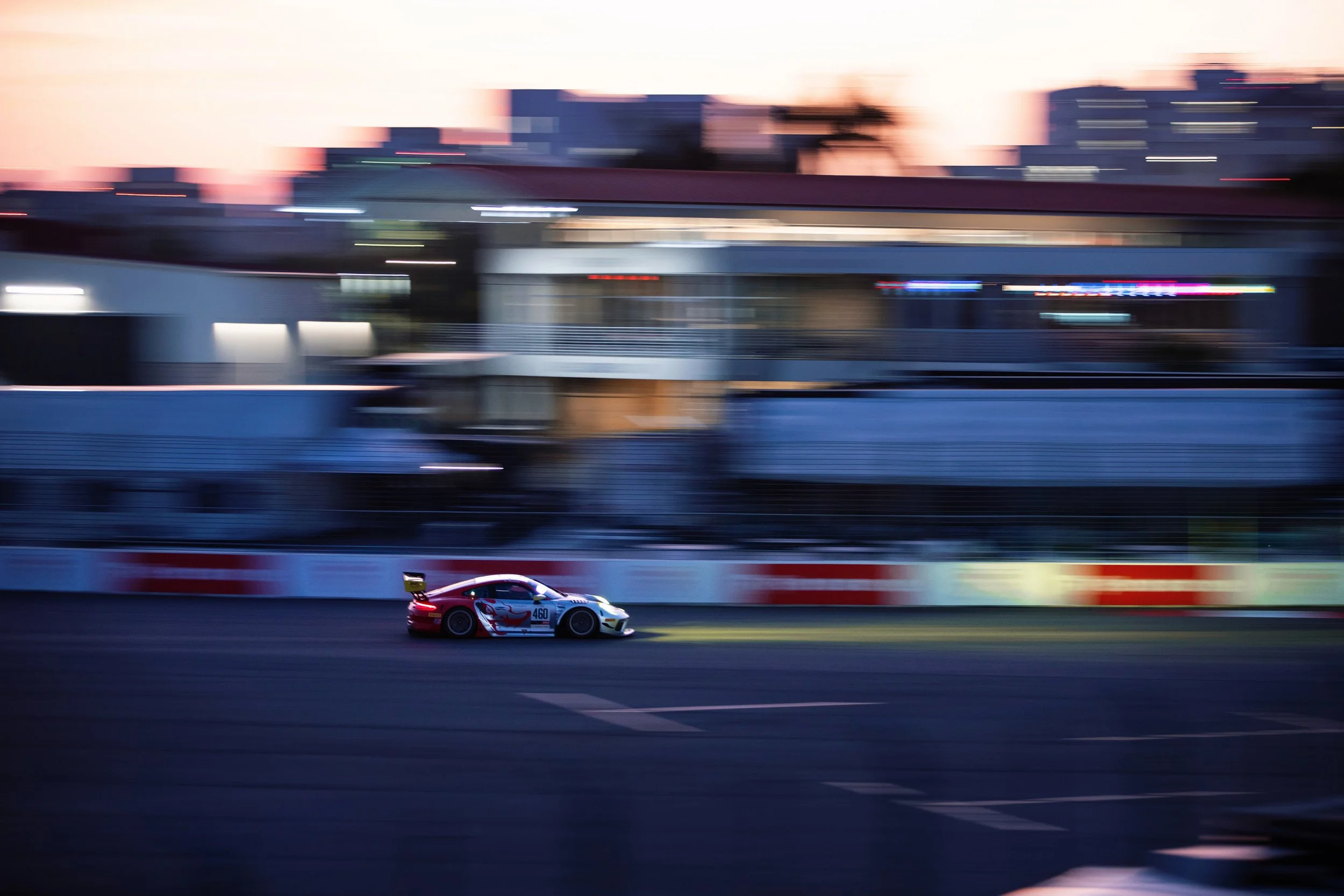 Cinematic automotive racing photography of a Porsche GT race car at speed against an urban skyline at sunset.