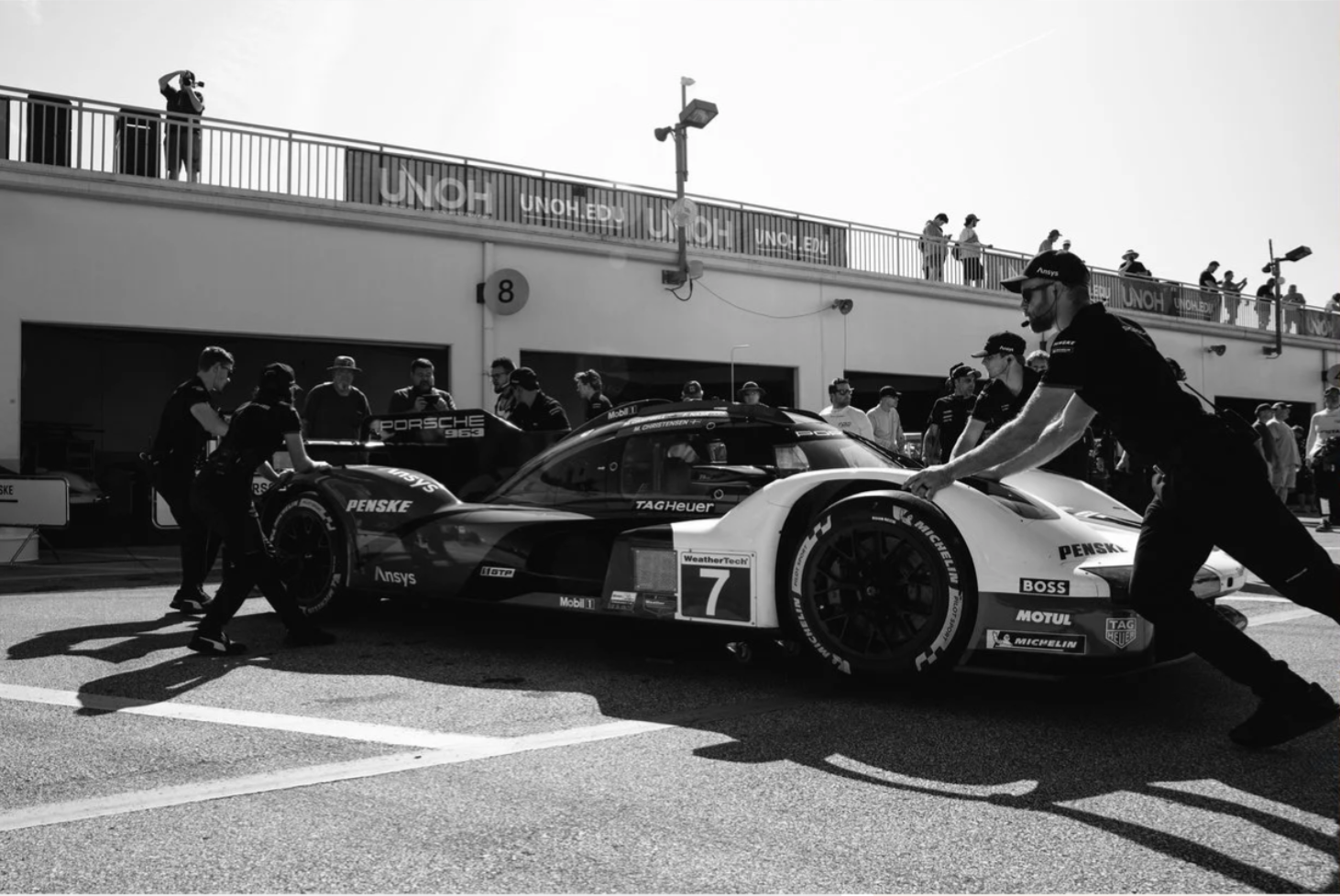 Black-and-white race weekend photography of Porsche Penske Motorsport crew members pushing a Porsche 963 race car through the IMSA pit lane.