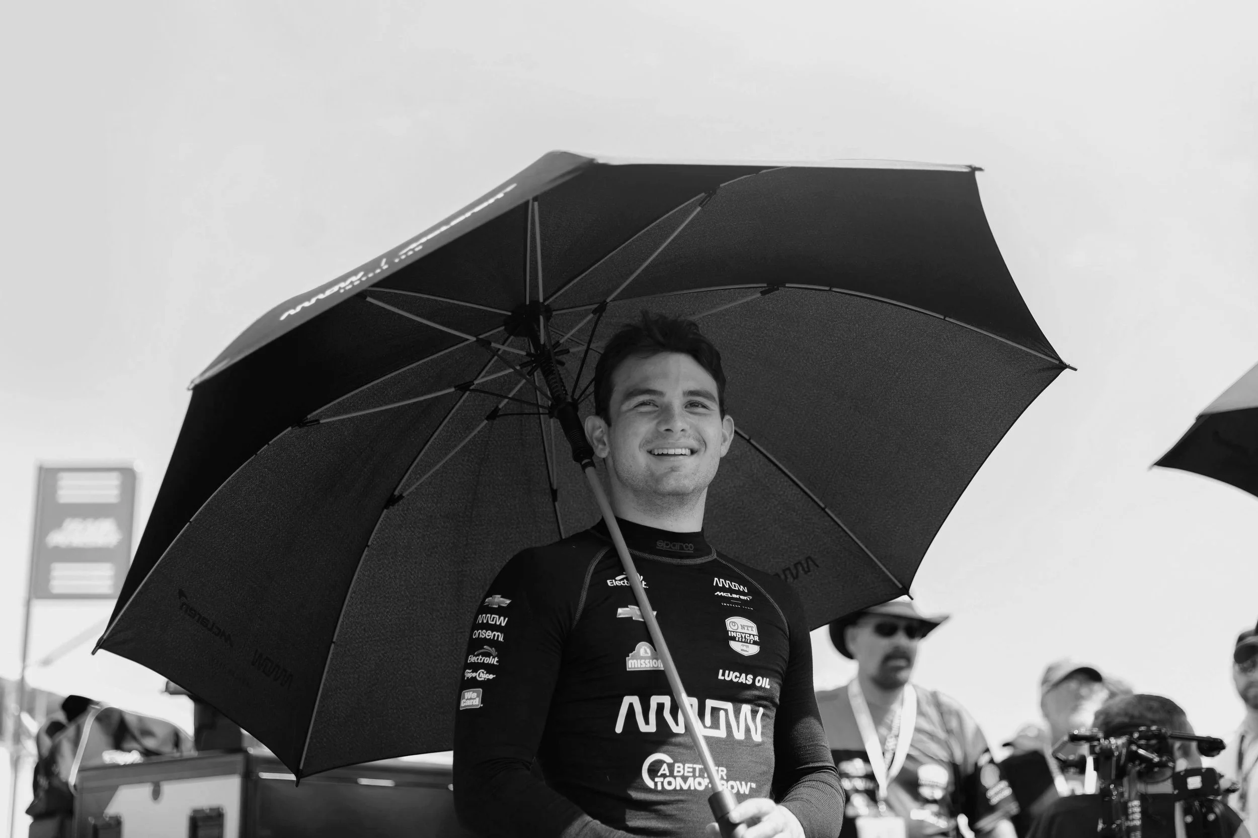 Black-and-white editorial motorsport portrait of Arrow McLaren IndyCar driver Pato O'Ward smiling beneath an umbrella during race weekend.