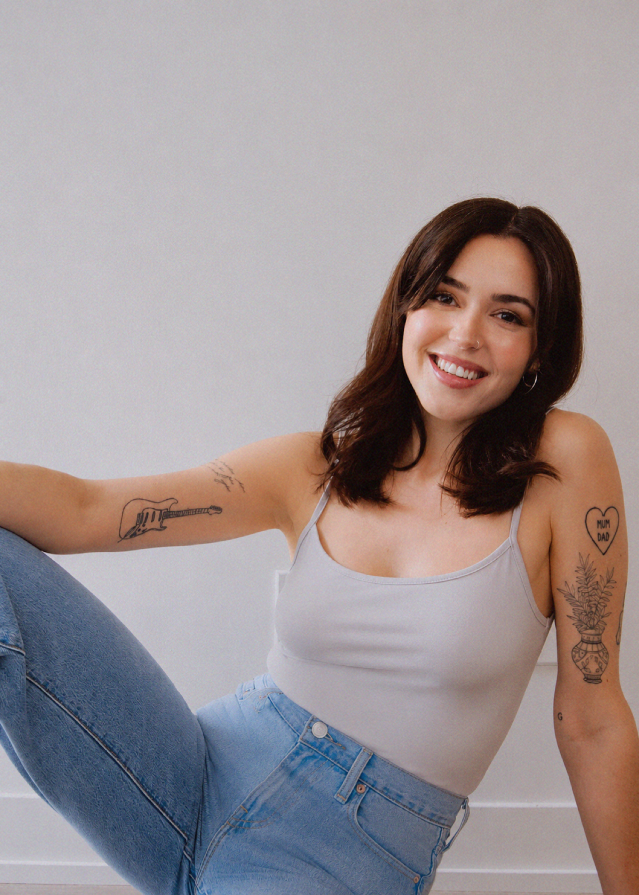 A young woman with shoulder-length dark hair and a nose piercing, smiling at the camera, sitting against a plain light-colored wall. She is wearing a white tank top and blue jeans, with tattoos on her arms.