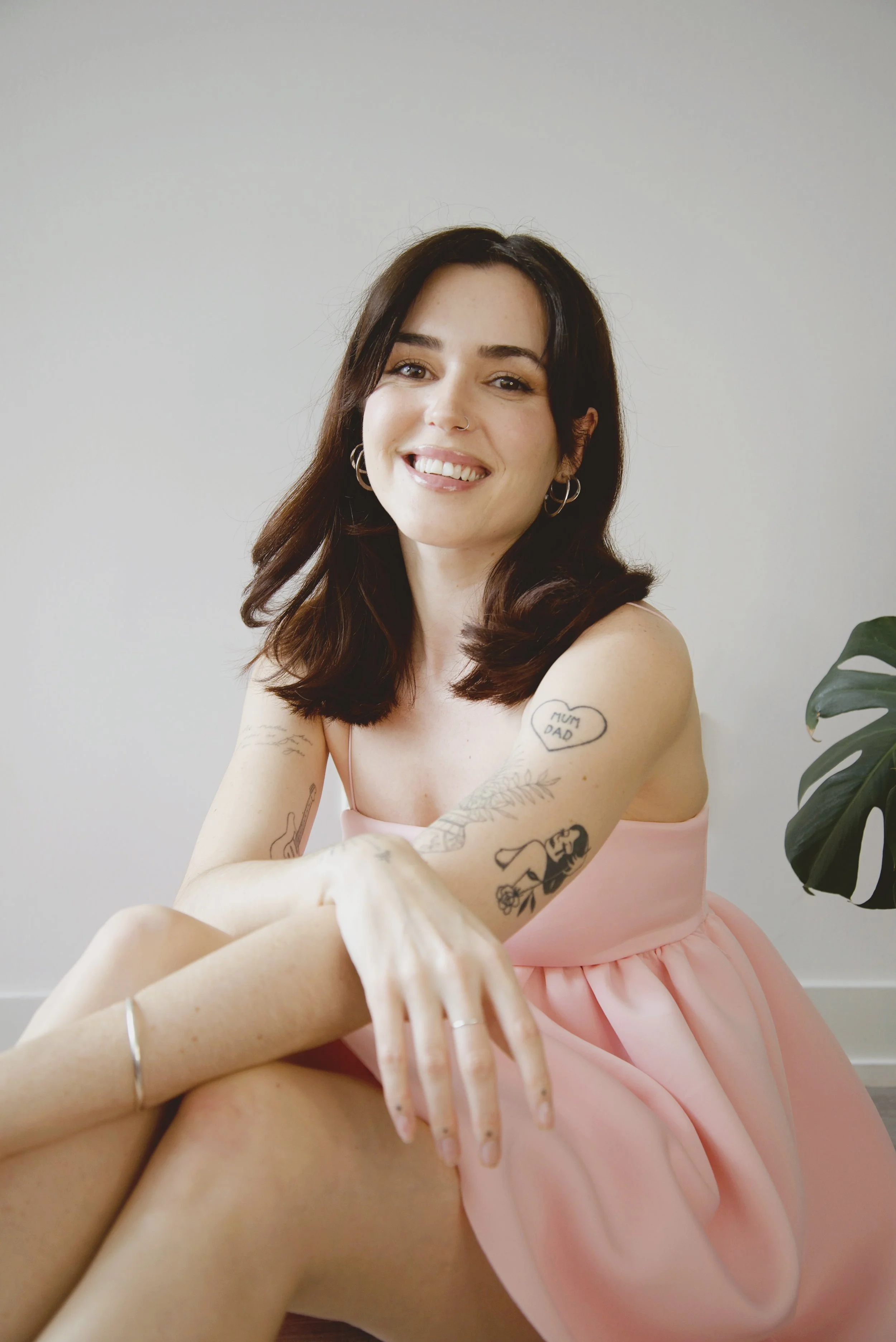 A woman in a pink dress smiling at the camera, with arms crossed and tattoos visible on her arms, sitting indoors with a plain light-colored wall and a plant nearby.