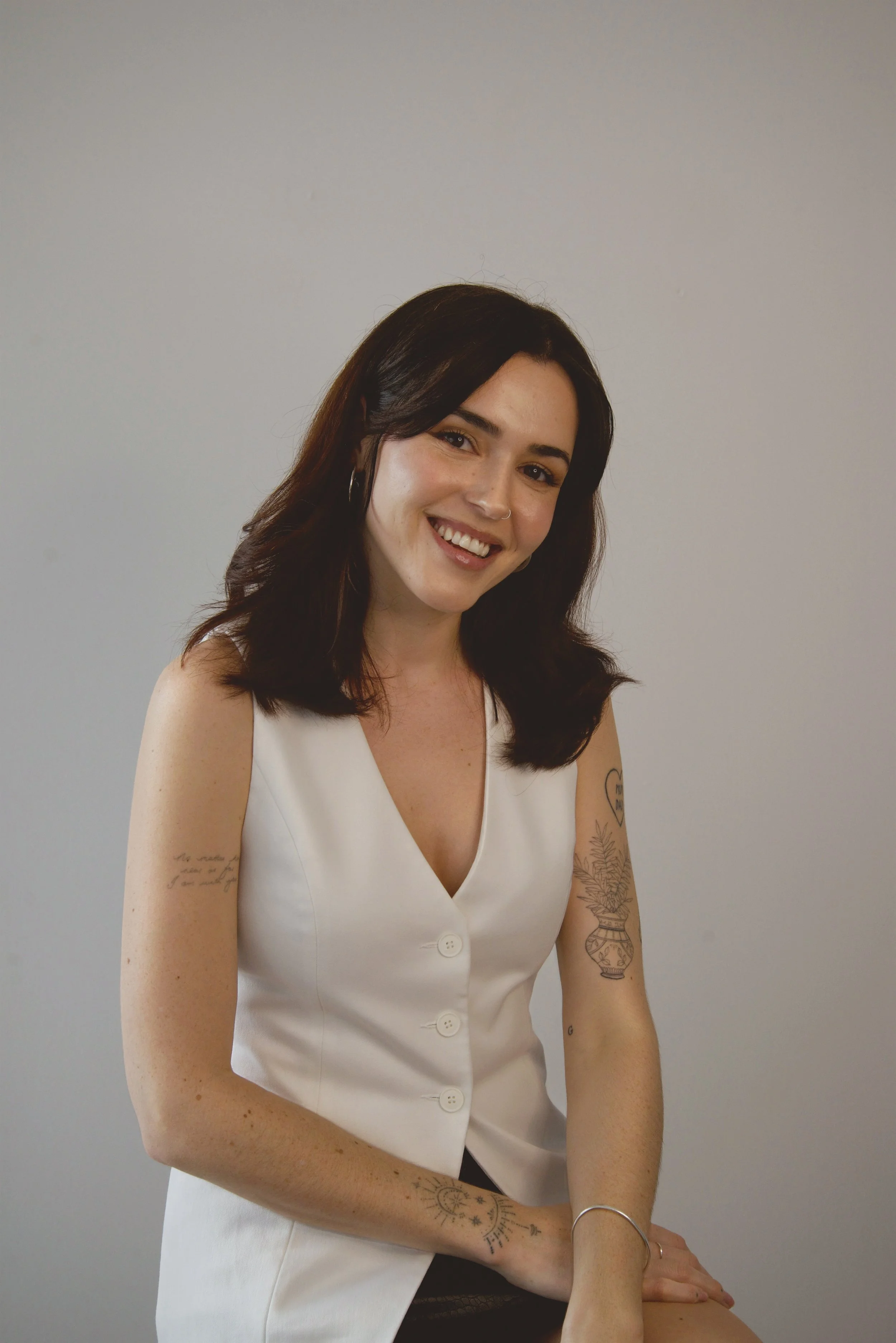 A young woman with dark brown hair, wearing a white sleeveless button-up dress, smiling with visible teeth, against a plain light gray background. She has tattoos on her left arm and shoulder, including a vase with flowers and some text on her right arm, and wears hoop earrings and a nose ring.