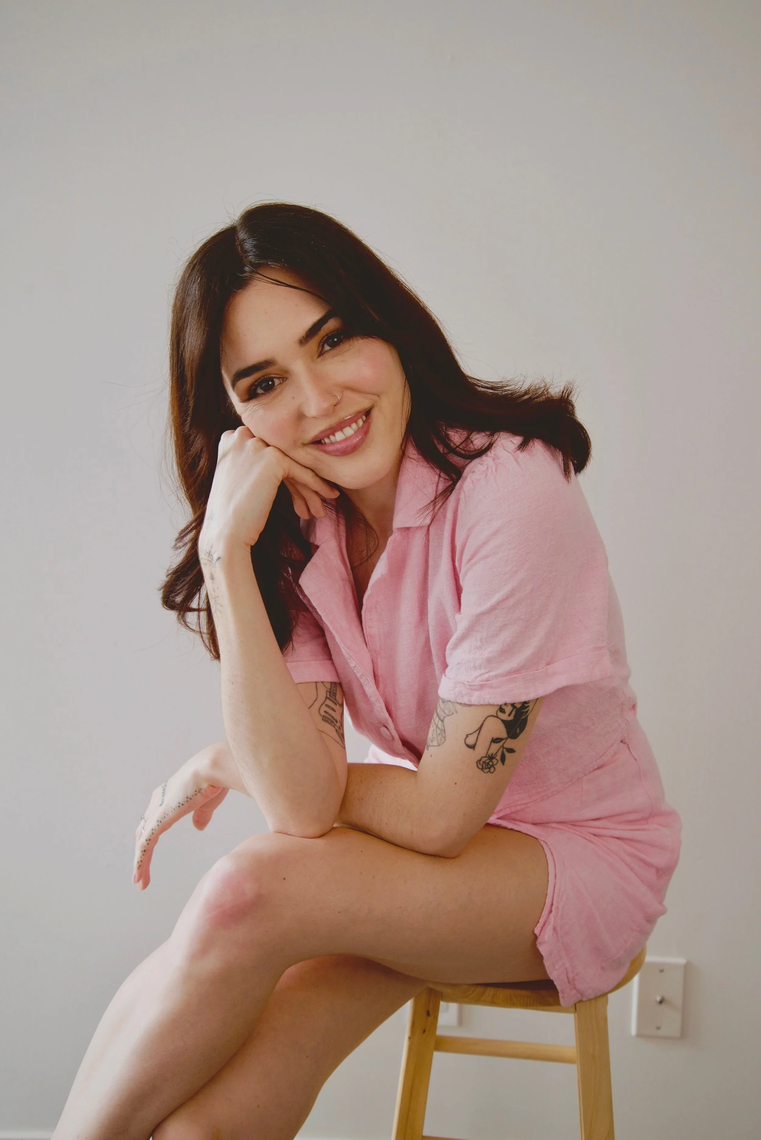 A smiling woman with dark hair and tattoos, wearing a pink shirt and shorts, sitting on a wooden stool against a plain white wall.