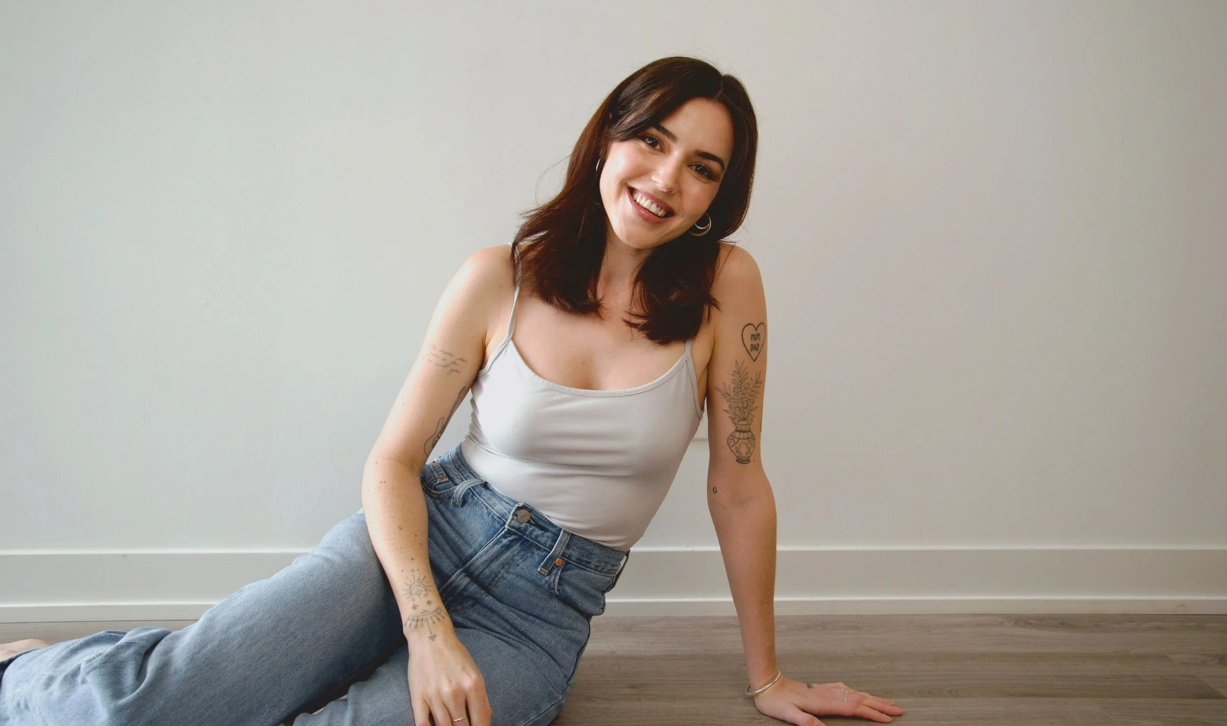 A young woman with dark brown hair sitting on the floor against a plain white wall, smiling at the camera. She is wearing a white tank top and blue jeans, with visible tattoos on her arms.