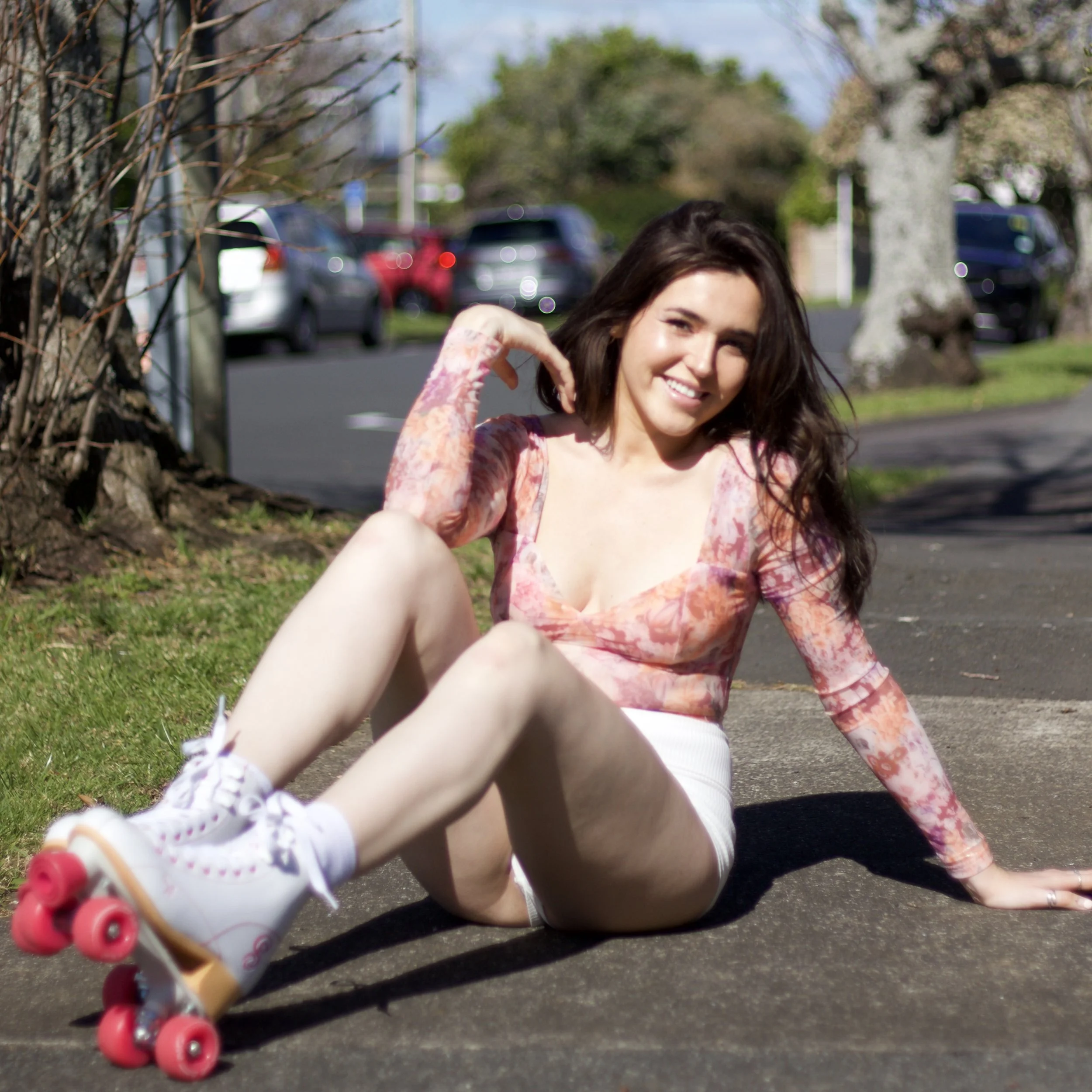 A young woman with dark hair smiling while sitting on the ground in a park, wearing a pink floral top and roller skates with pink wheels, with a background of cars and trees.