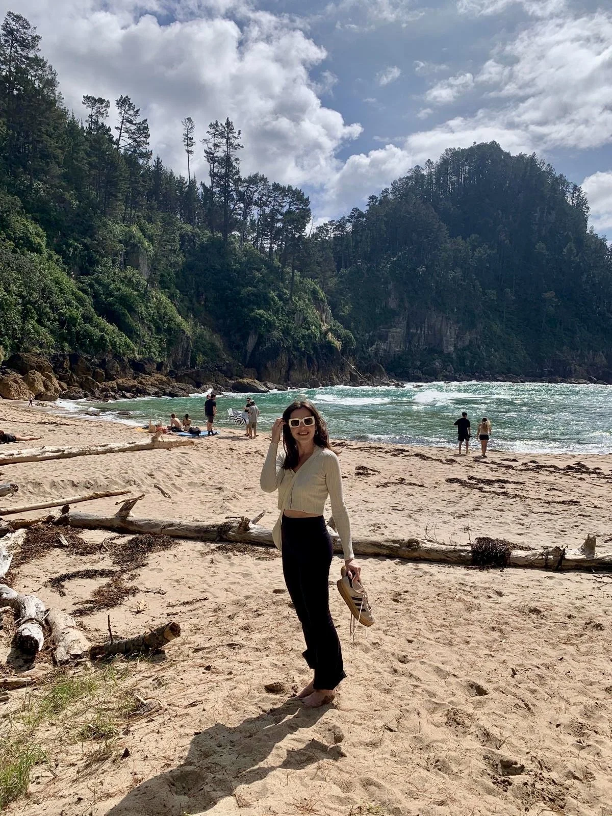 Woman standing on a sandy beach, smiling, holding shoes, with ocean and cliffs in background, under partly cloudy sky.