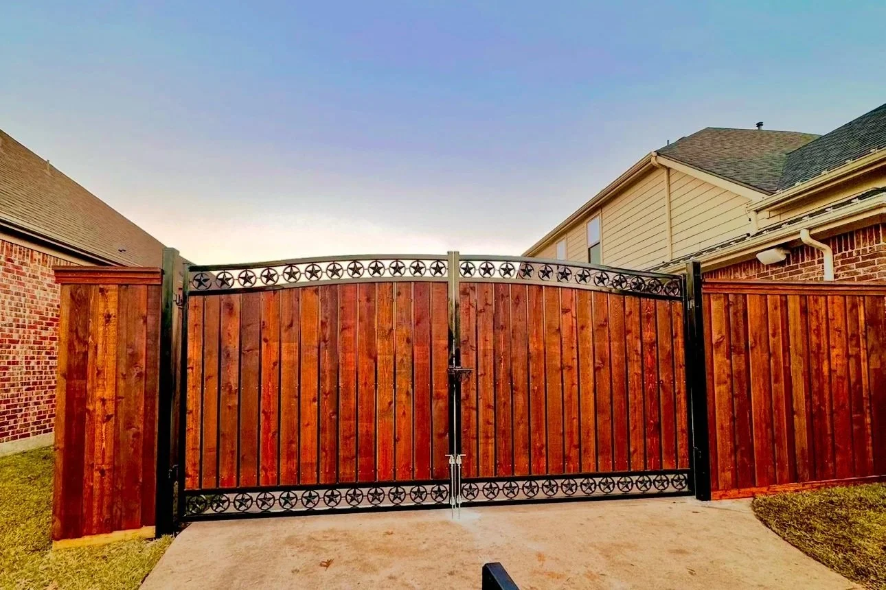 A wooden gate with decorative metal top and bottom panels, set between brick and wooden fences on either side, with a concrete pavement in front and houses with sloped roofs in the background under a clear sky.