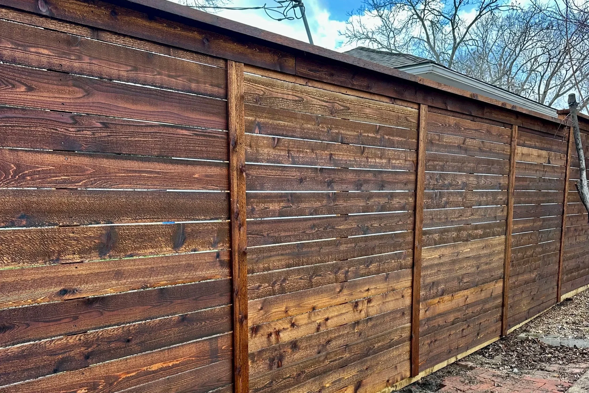 A newly built wooden fence with stained horizontal planks and vertical support beams, with a house and trees visible in the background under a blue sky with some clouds.