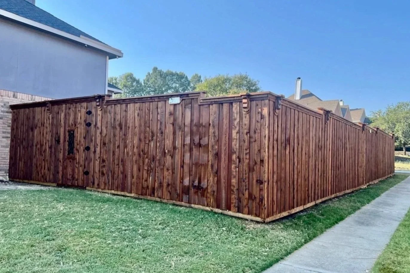Wooden privacy fence enclosing a backyard near a sidewalk, with a corner of a house visible on the left and trees in the background.