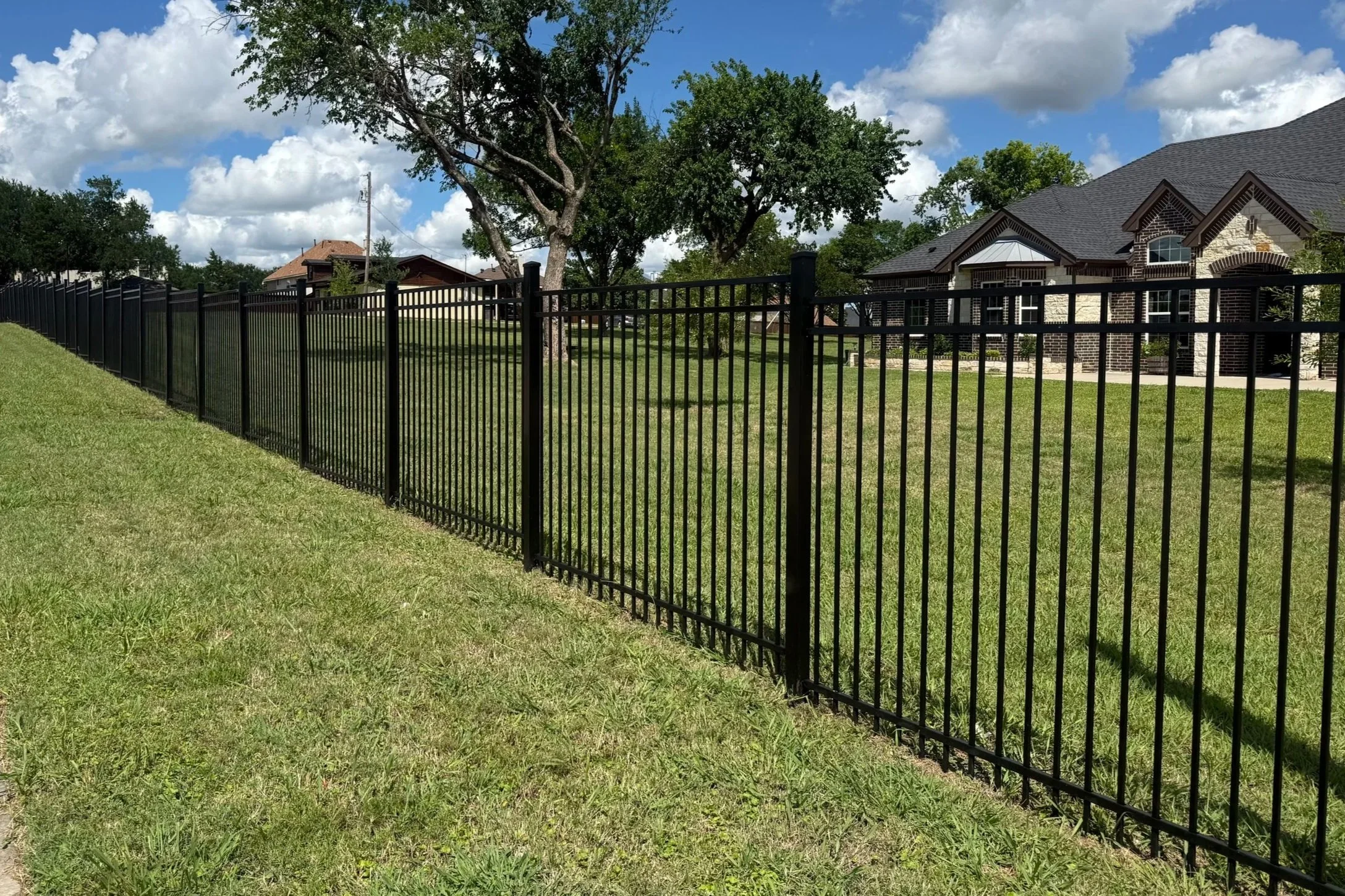 A black metal fence runs along a green lawn in a residential backyard. In the background, there are trees with full green leaves, and a house with a brick and stone exterior. The sky is partly cloudy with patches of blue sky visible.