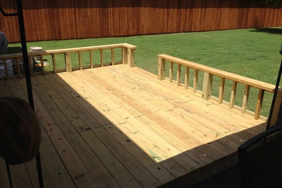 A newly built wooden deck in a backyard, partially shaded, with a wooden fence and green grass in the background.