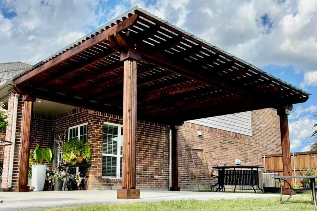 A wooden patio cover attached to a brick house, with outdoor furniture and potted plants underneath, surrounded by a grassy yard and a blue sky with clouds.
