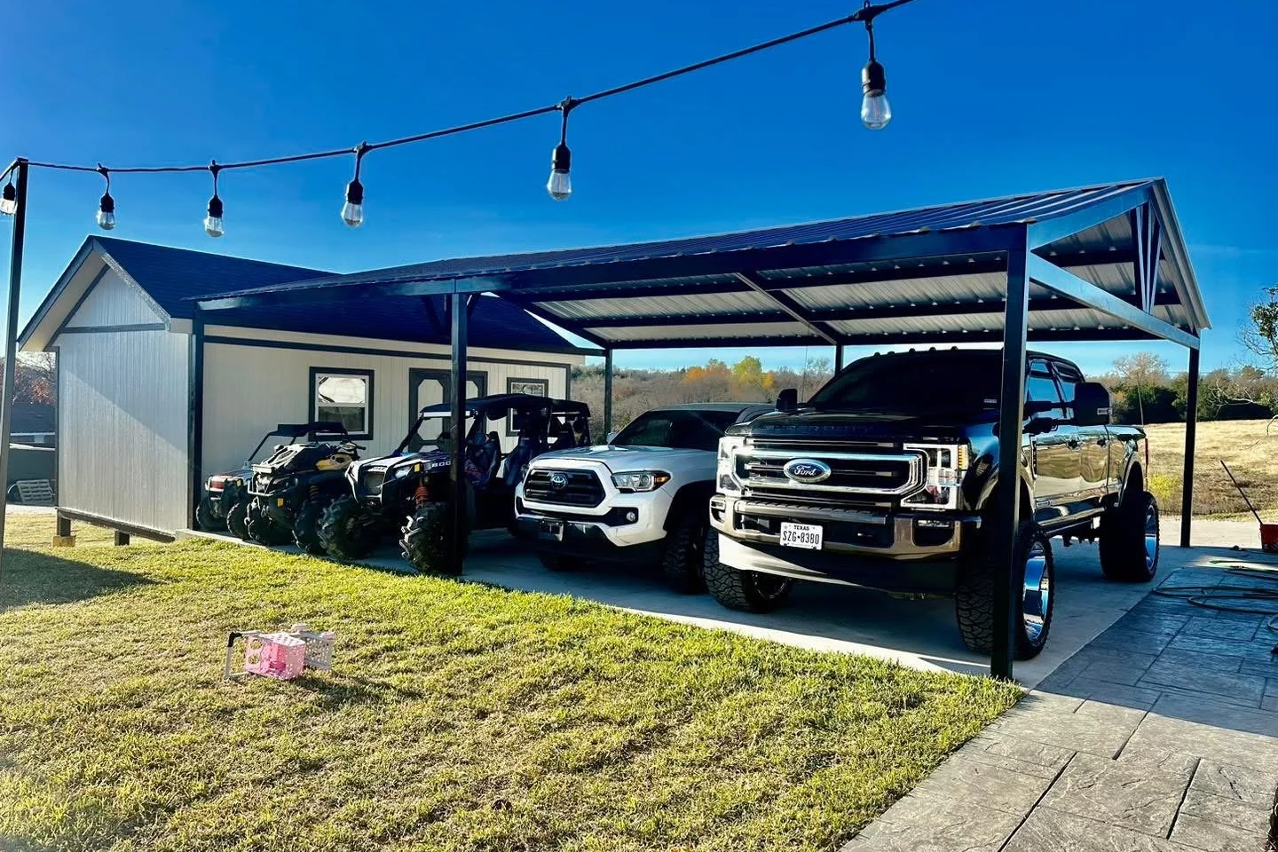 A metal carport with three vehicles, including a Ford pickup truck, an SUV, and a large utility vehicle, parked on a concrete driveway beside a small white building. String lights are hanging overhead, and a patch of grass with a pink toy is in the foreground.