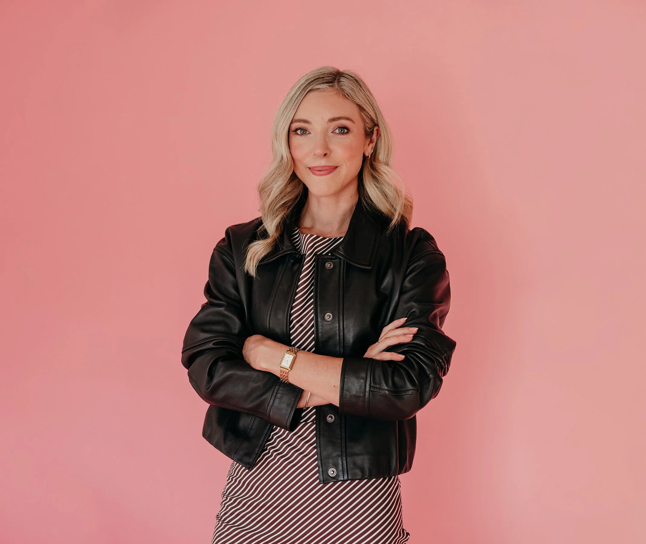 Chelsea, the founder, is a woman with blonde hair styled in loose waves, wearing a black leather jacket over a striped dress, standing against a pink background.