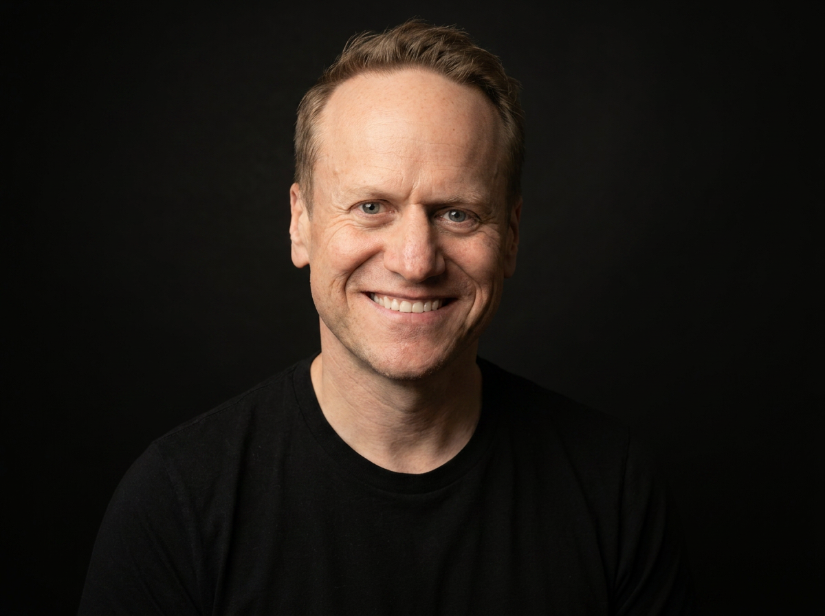 Portrait of a smiling man with short blonde hair wearing a black shirt against a dark background.