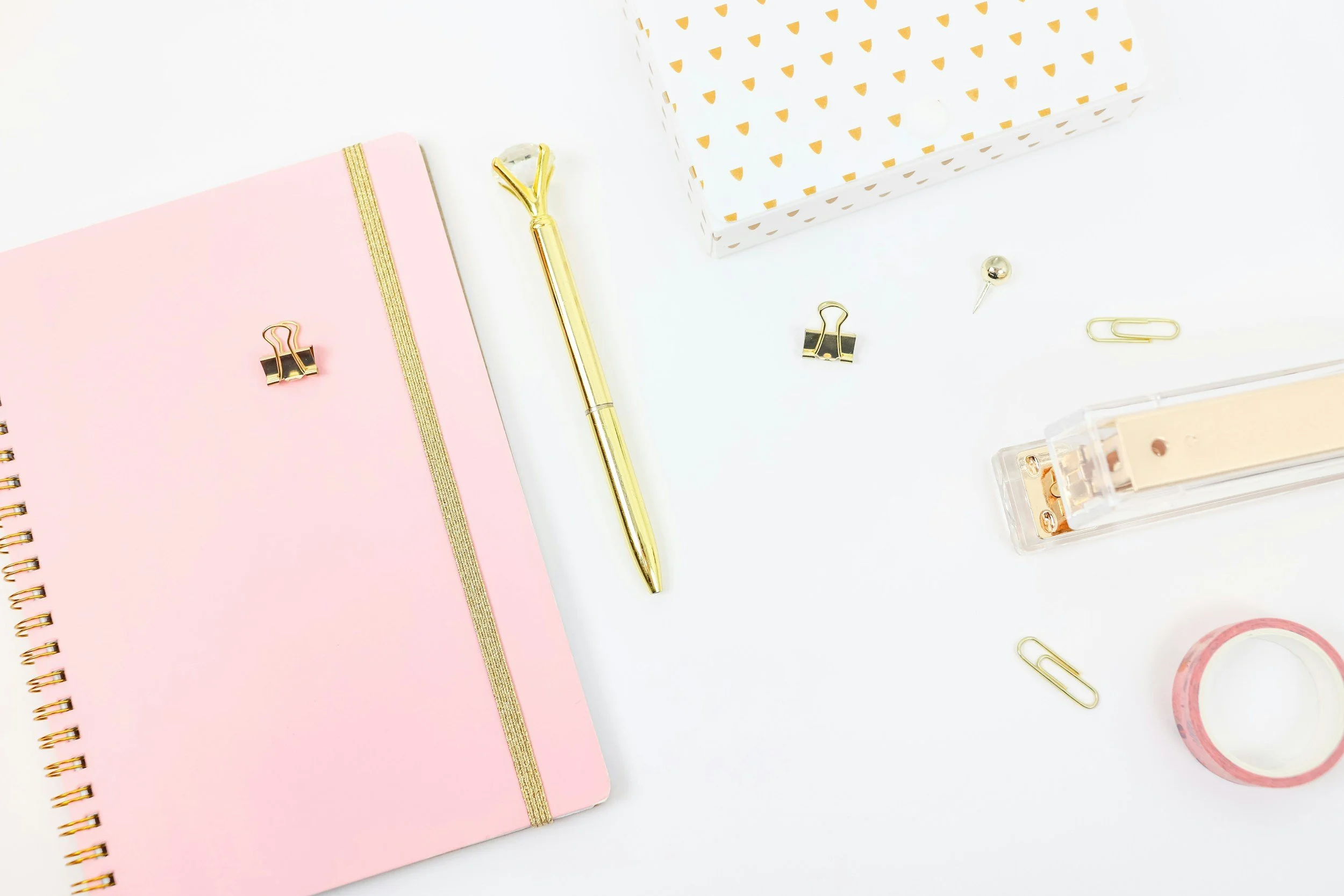 Cuaderno rosa con una banda dorada en la portada, una pluma dorada, clips de papel dorados, cinta adhesiva rosa y cinta de doble cara en un fondo blanco con una caja decorativa de fondo.