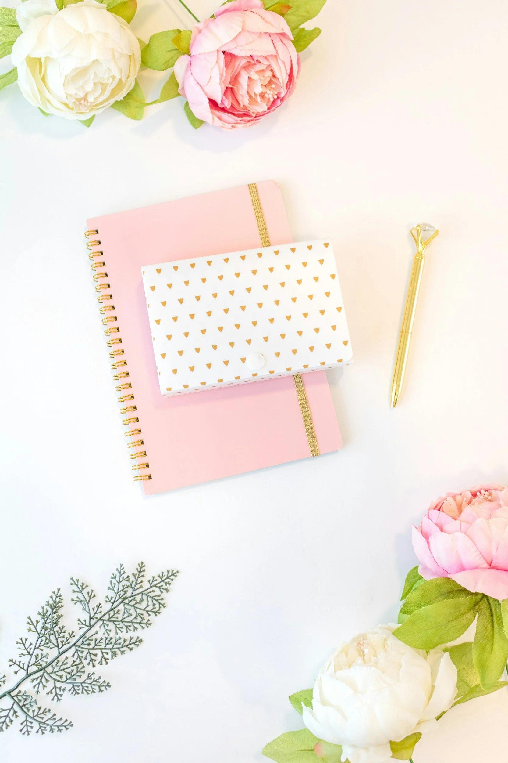 Cuatro flores grandes, dos blancas y dos rosas, y varias hojas verdes, junto a libretas de color rosa y un bolígrafo dorado en una mesa blanca, decoraciones florales adicionales.