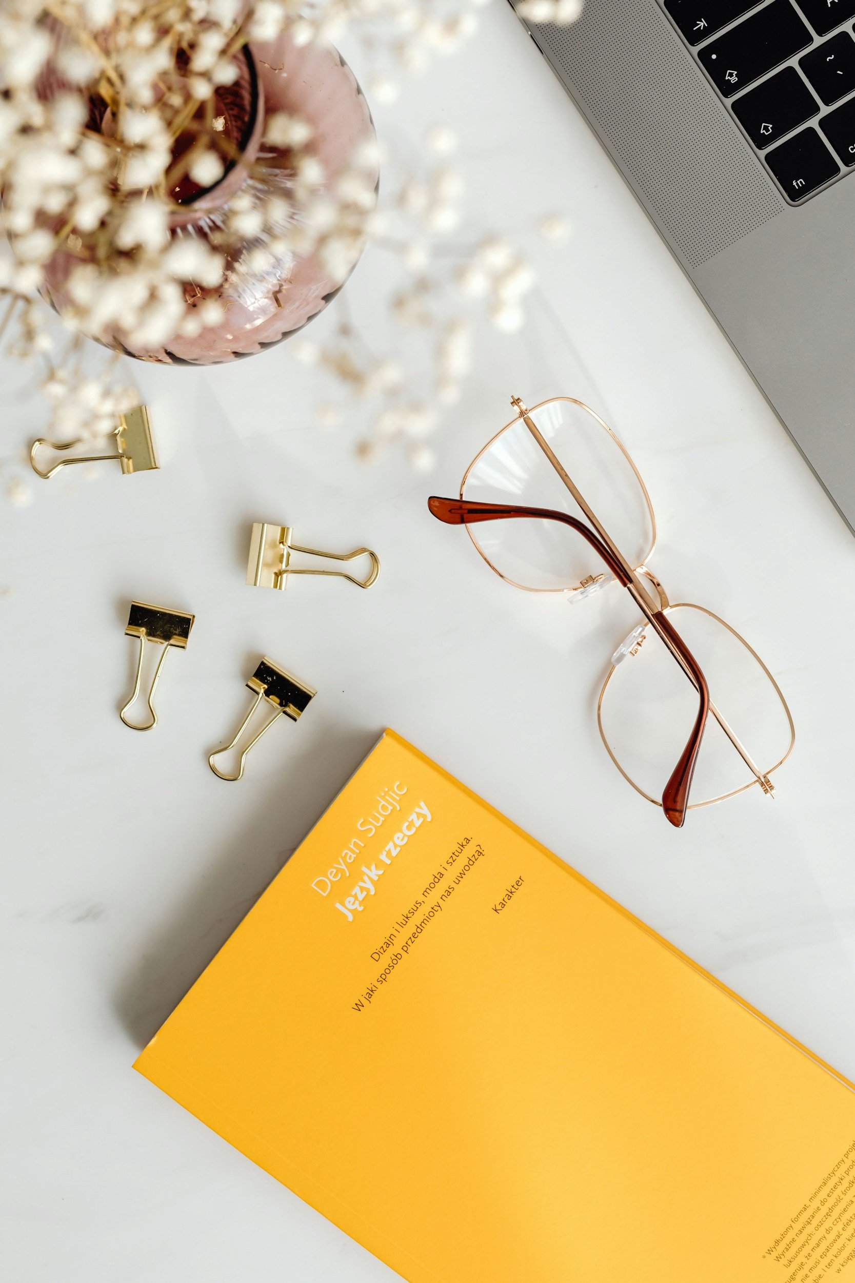 Vista superior de un escritorio con gafas de montura metálica color rosa, varios clips dorados, una maceta con flores blancas, un libro de color amarillo y una laptop plateada.