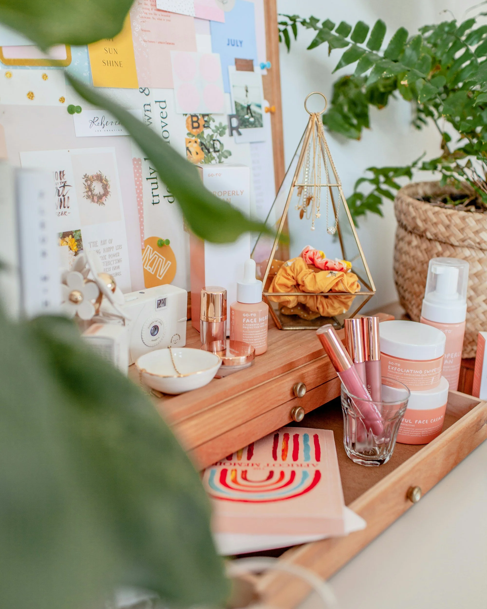 Organizador de escritorio de madera con varios productos de cuidado personal, algunos envases y una decoración con joyas colgando. En el fondo, un tablero de notas y plantas verdes en una maceta de mimbre.