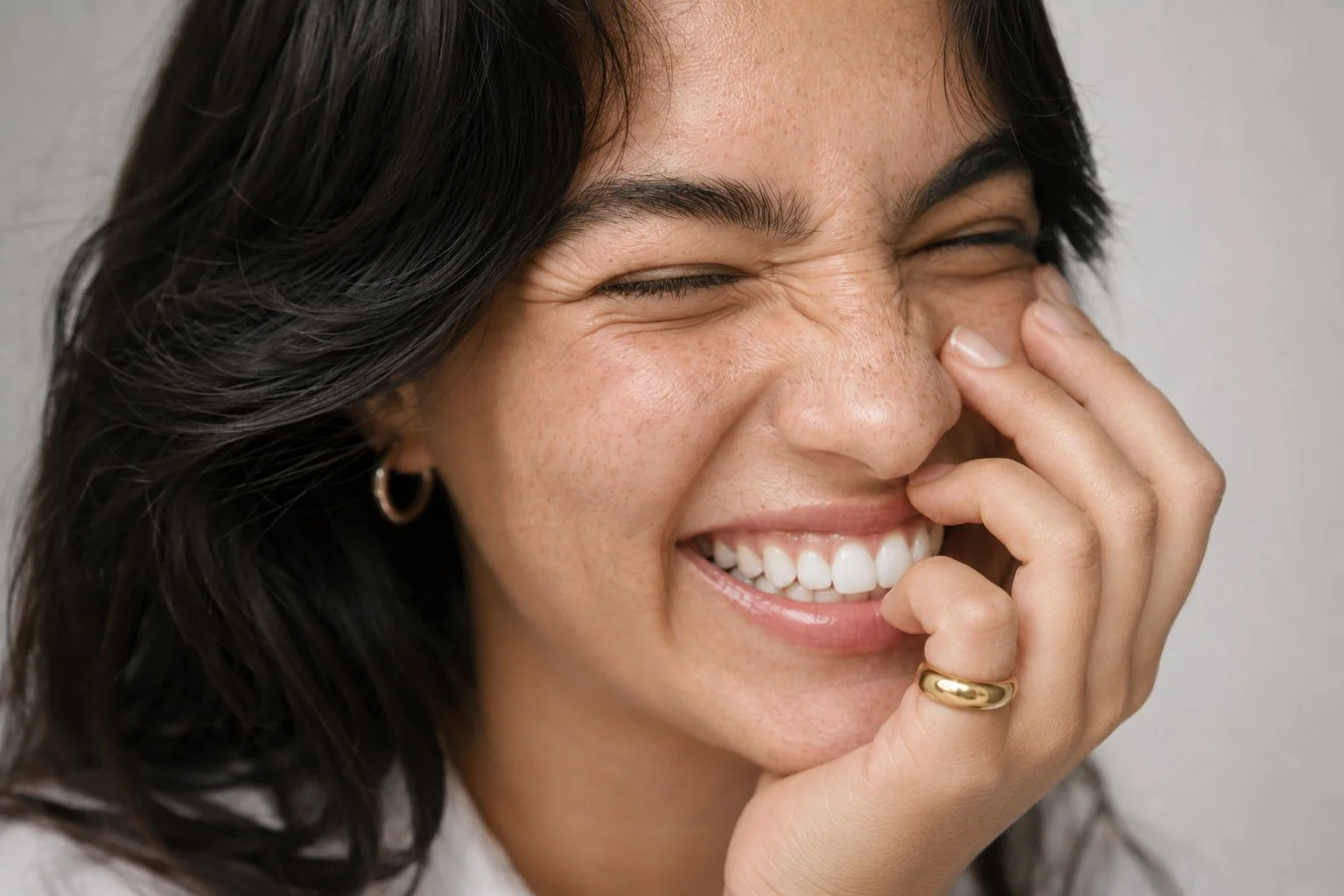 A woman with dark hair laughing and covering her face with her hand, showing a big smile with white teeth.