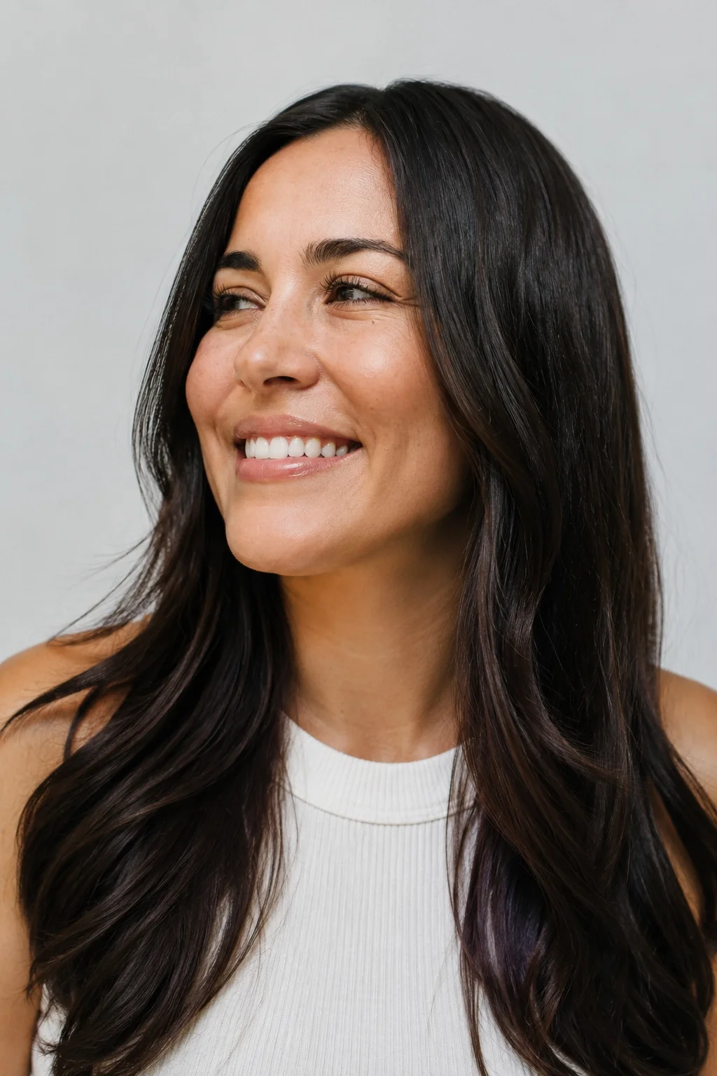 A woman smiling and looking to the side, with long dark hair and a white sleeveless top.