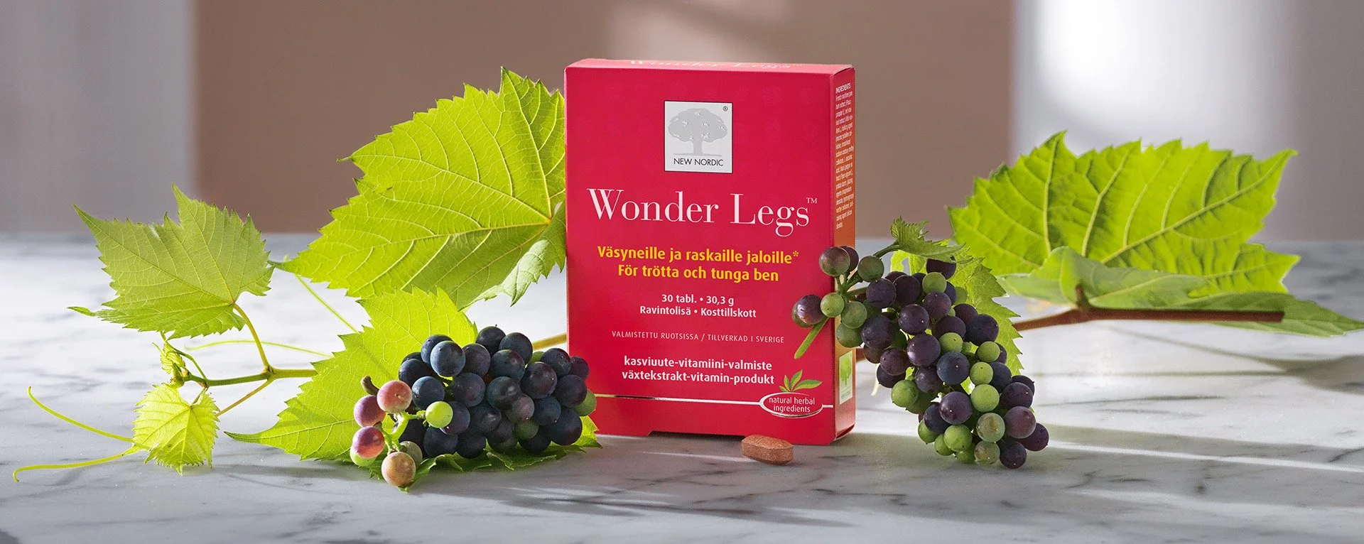 Punainen lääkelaatikko, jonka nimi on Wonder Legs, on asetettu valkoisen ja harmahtavan taustan edessä. Laatikko on koristeltu vihreillä lehdillä ja rypäleillä, jotka ympäröivät sitä.