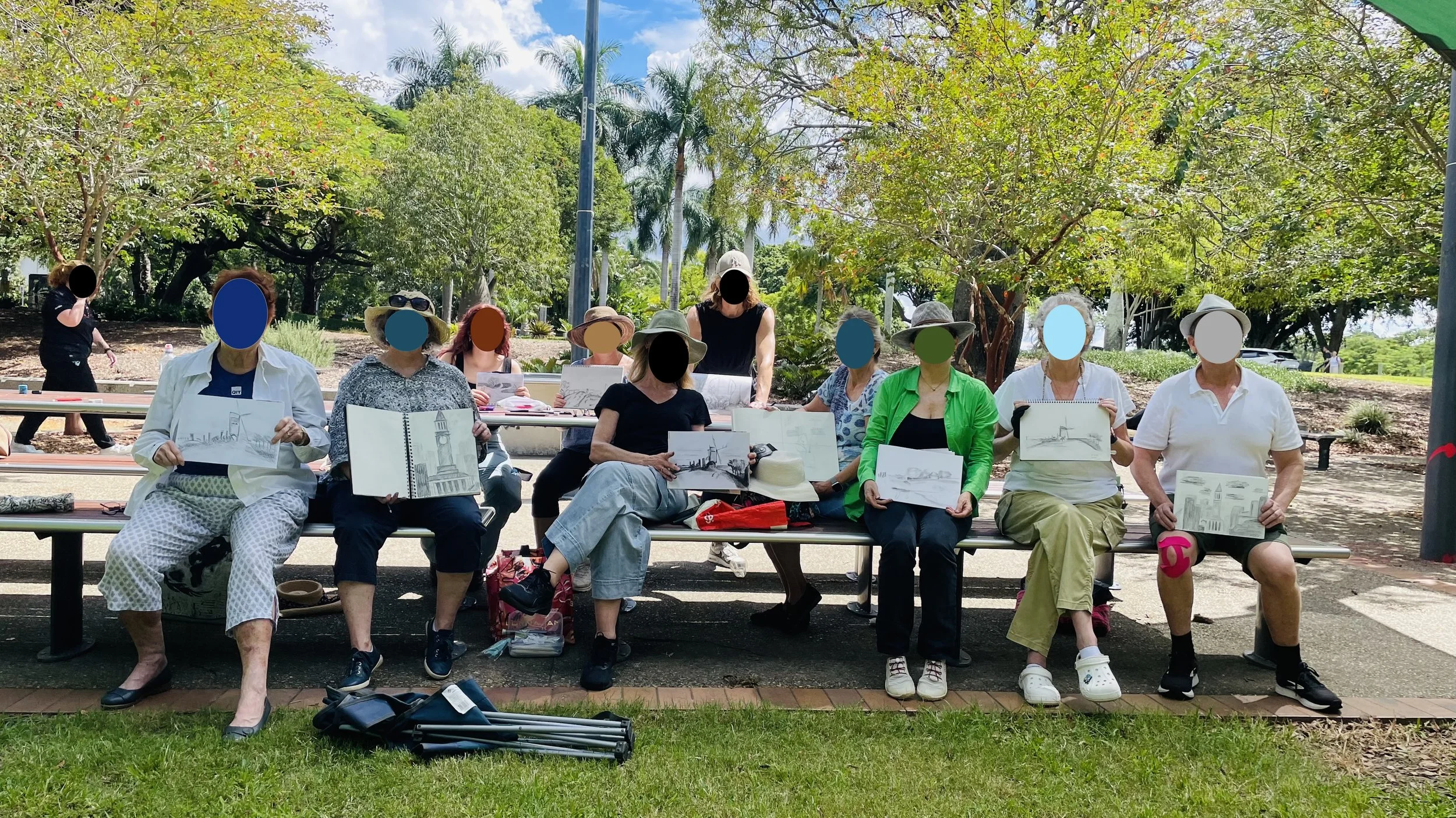 A group of predominantly older women and a couple of men sitting on benches outdoors, holding up sketchbooks or drawings. Some are wearing hats and sunglasses. The scene is set in a park with trees and greenery in the background, and a partly cloudy sky.