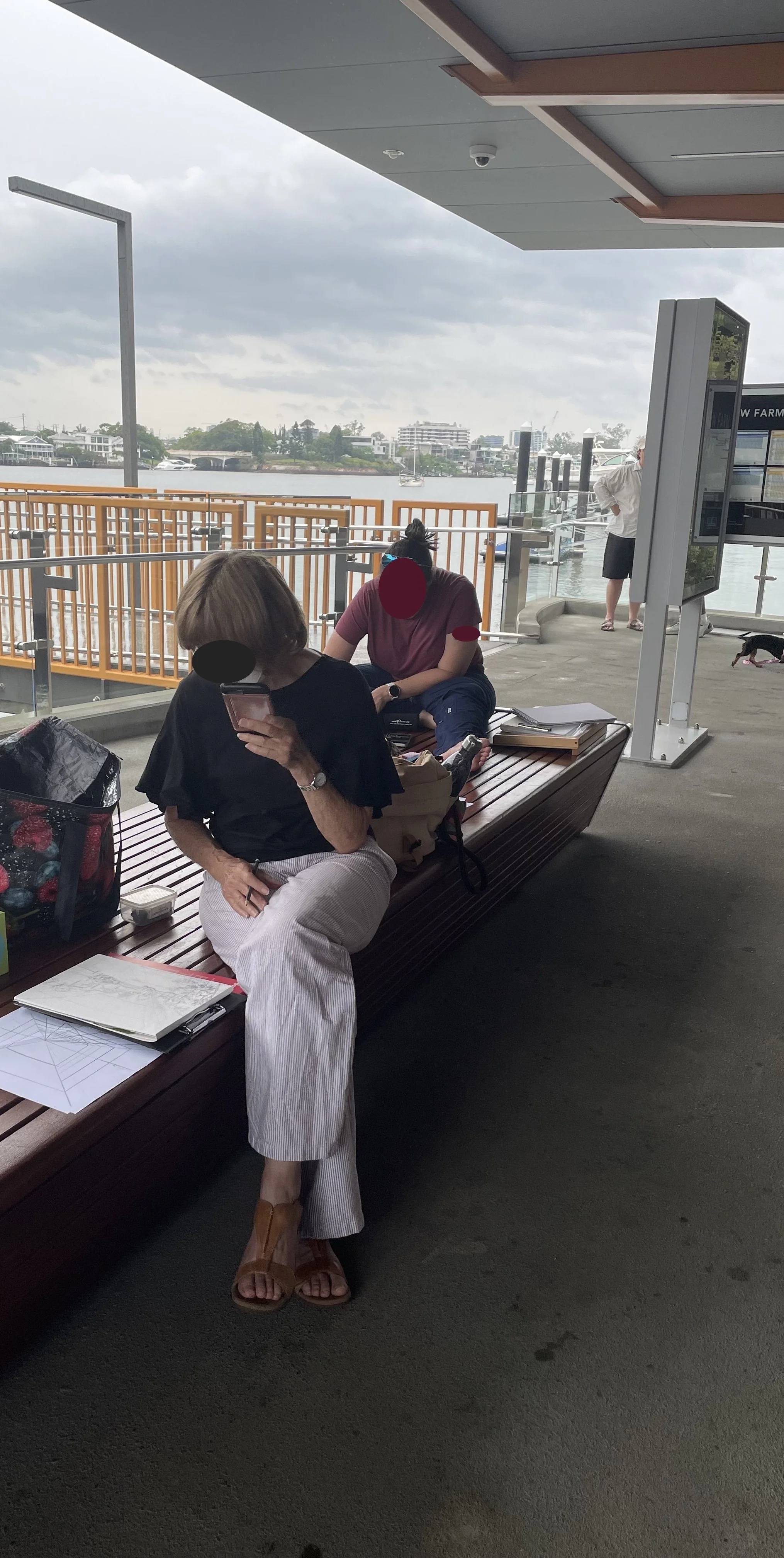 People sitting on a bench near a waterway, with a woman looking at her phone and sketching and another person sitting with a laptop and papers. There is an informational sign and a man in the background.