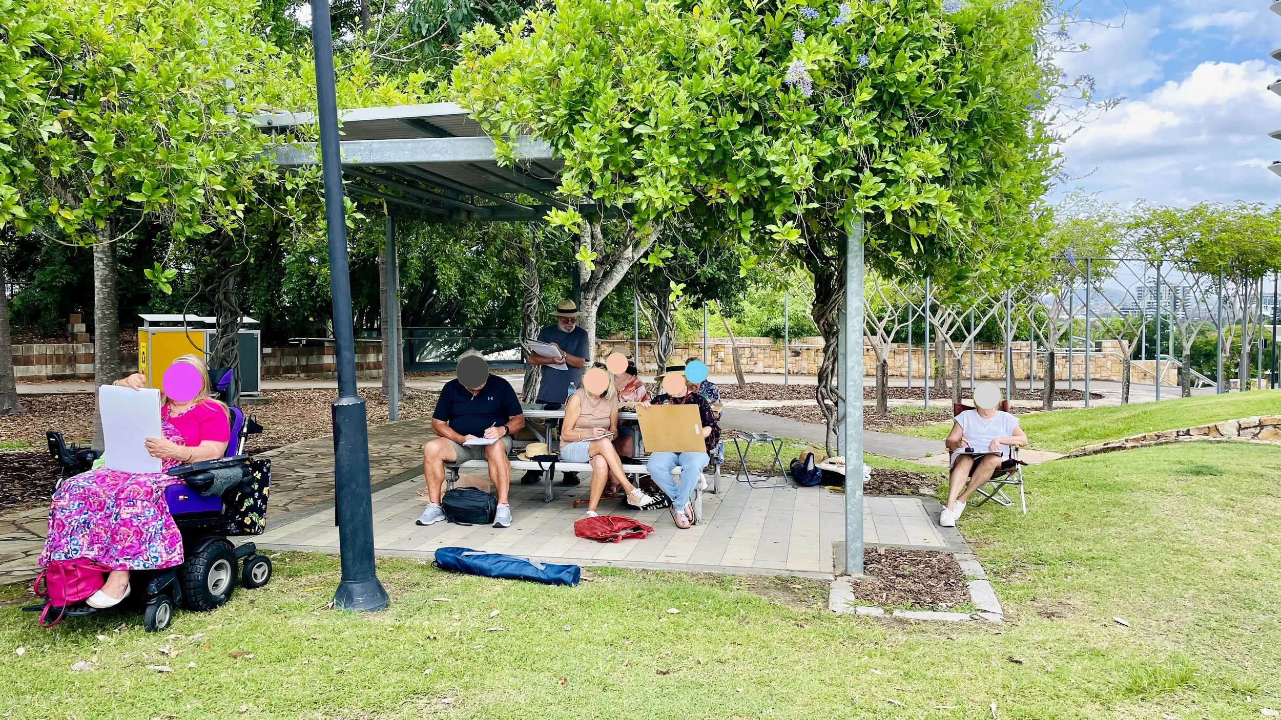 Group of people sitting on benches and chairs under a shaded area with trees, sketching and painting, with a cityscape in the background.