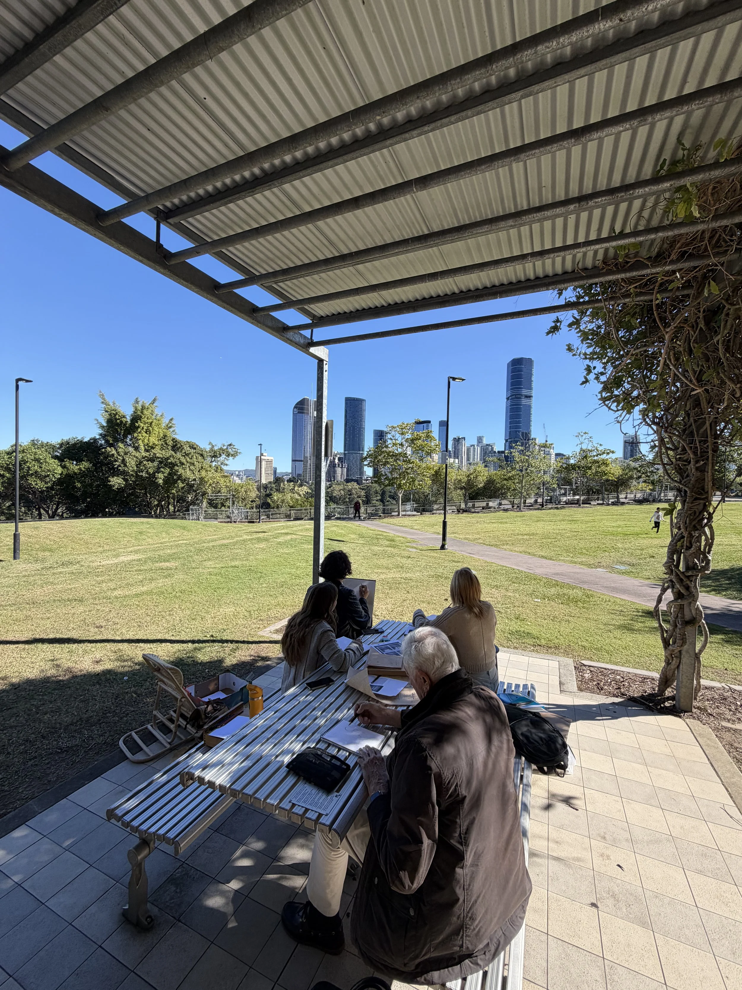 People sitting at a picnic table sketching  under a canopy in a park, with a city skyline in the background on a sunny day.