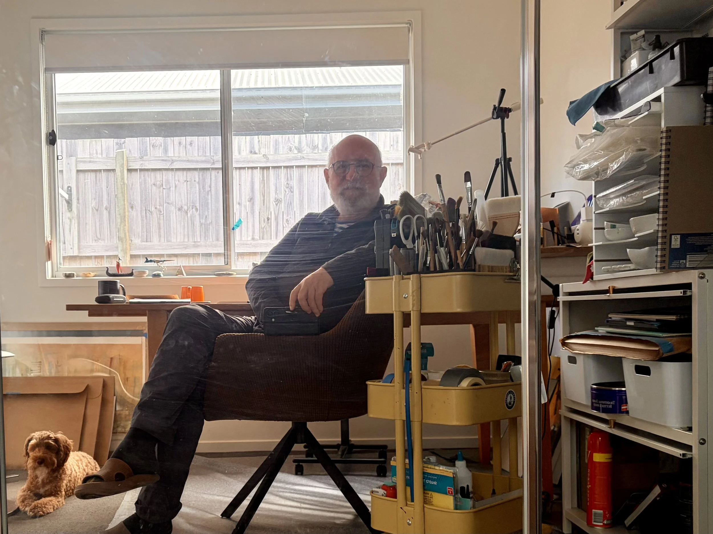A man with glasses and a beard sitting at a desk in a room filled with various objects, with a window showing an outside wooden fence, and a small brown dog sitting on the floor nearby.