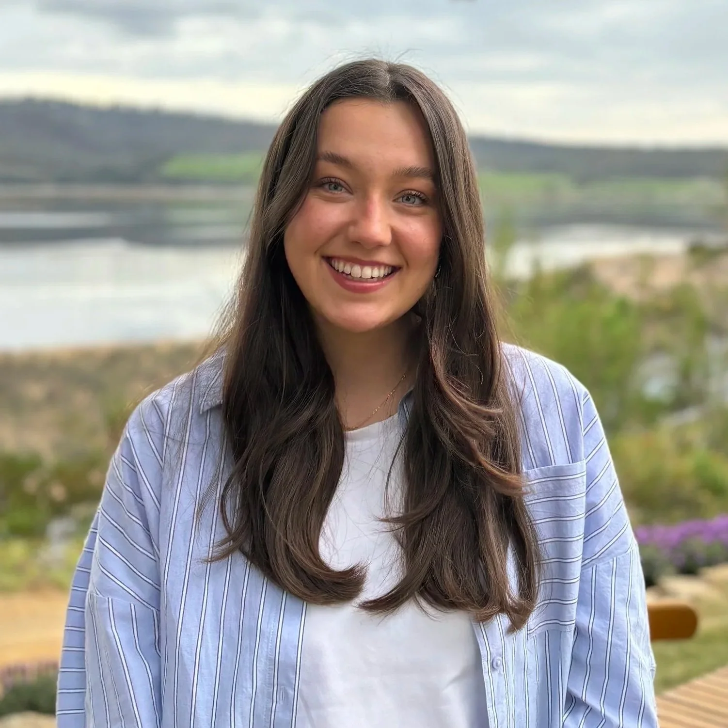 A young woman with long brown hair and blue eyes smiling outdoors with a body of water, green hills, and cloudy sky in the background.
