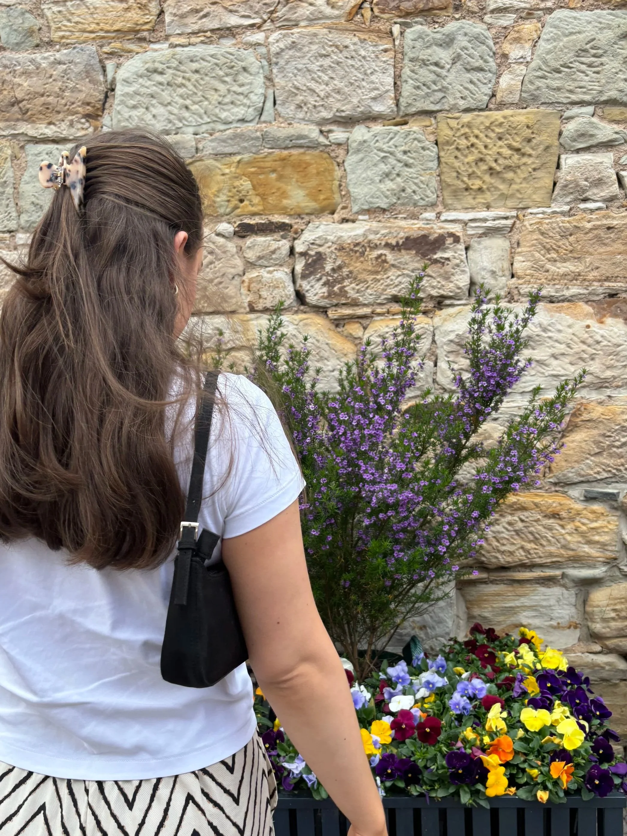 A woman with long brown hair in a beige and black hair clip, wearing a white shirt and patterned pants, stands near a stone wall with vibrant purple and yellow flowers.