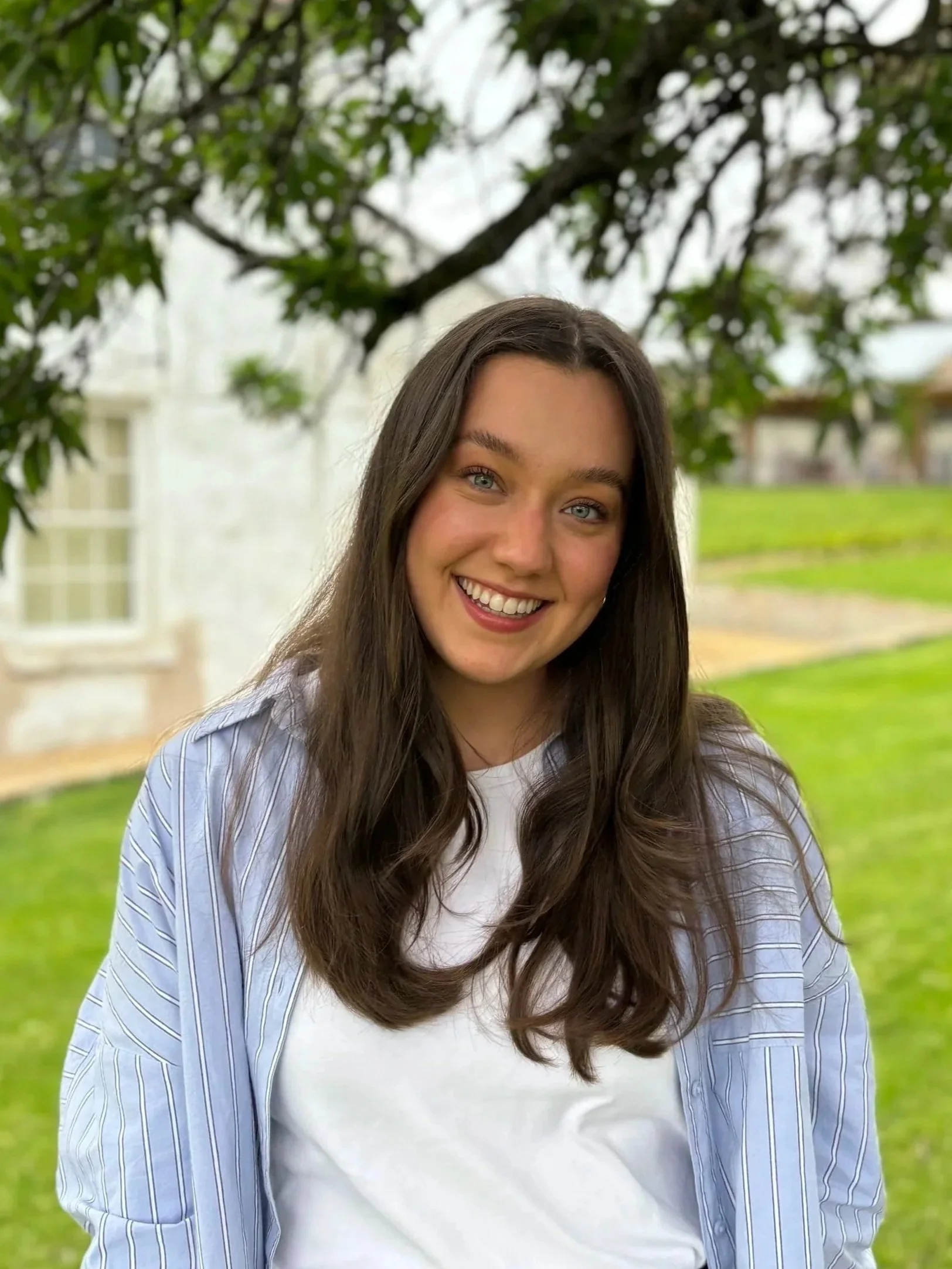 Smile of young woman with long brown hair, light blue eyes, wearing a white t-shirt and a blue striped shirt, standing outdoors with greenery and trees in the background.