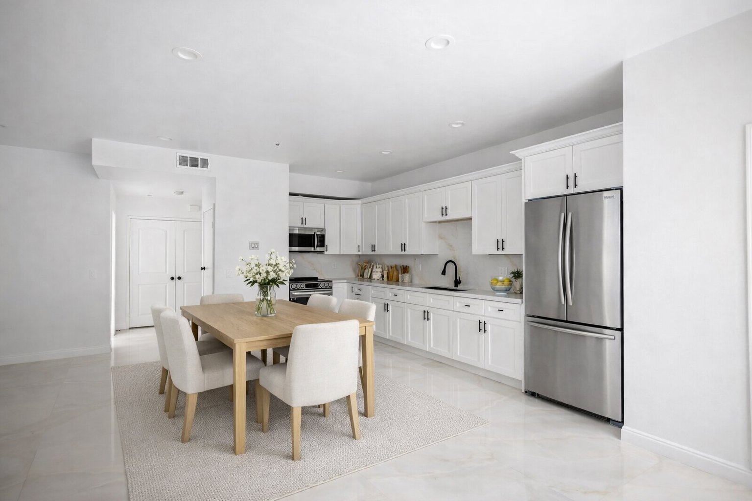 Modern kitchen with white cabinets, stainless steel refrigerator, black faucet, and a dining table with white chairs and a vase of flowers.