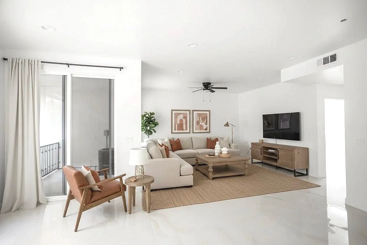 Bright living room with white walls, beige sectional sofa, wooden armchair, glass sliding doors with white curtains, flat-screen TV on wooden console, coffee table, houseplant, framed artwork, ceiling fan, and beige area rug.
