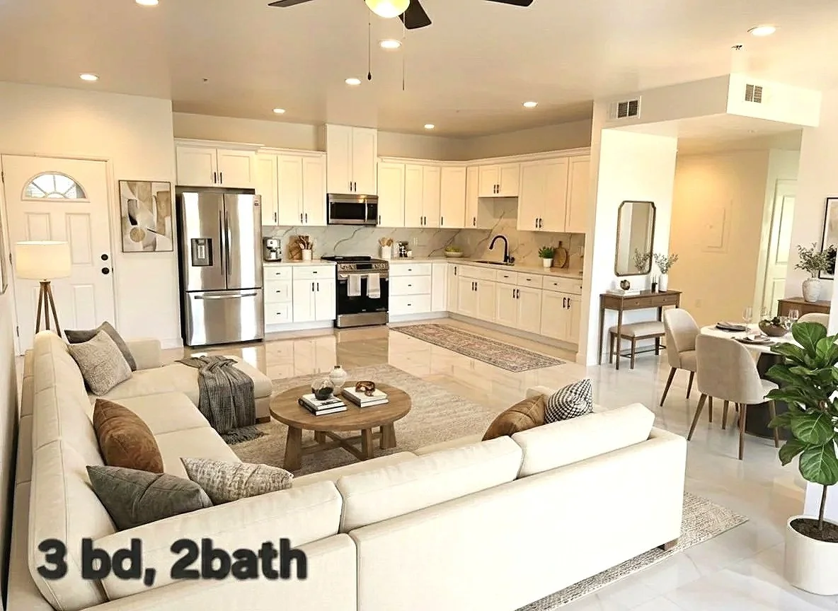 Open-concept living room and kitchen with white cabinets, stainless steel appliances, and a small dining table with four chairs. The living room has a large white sofa, a round coffee table, and decorative pillows. There is a painting on the wall near the door, and potted plants near the seating area.
