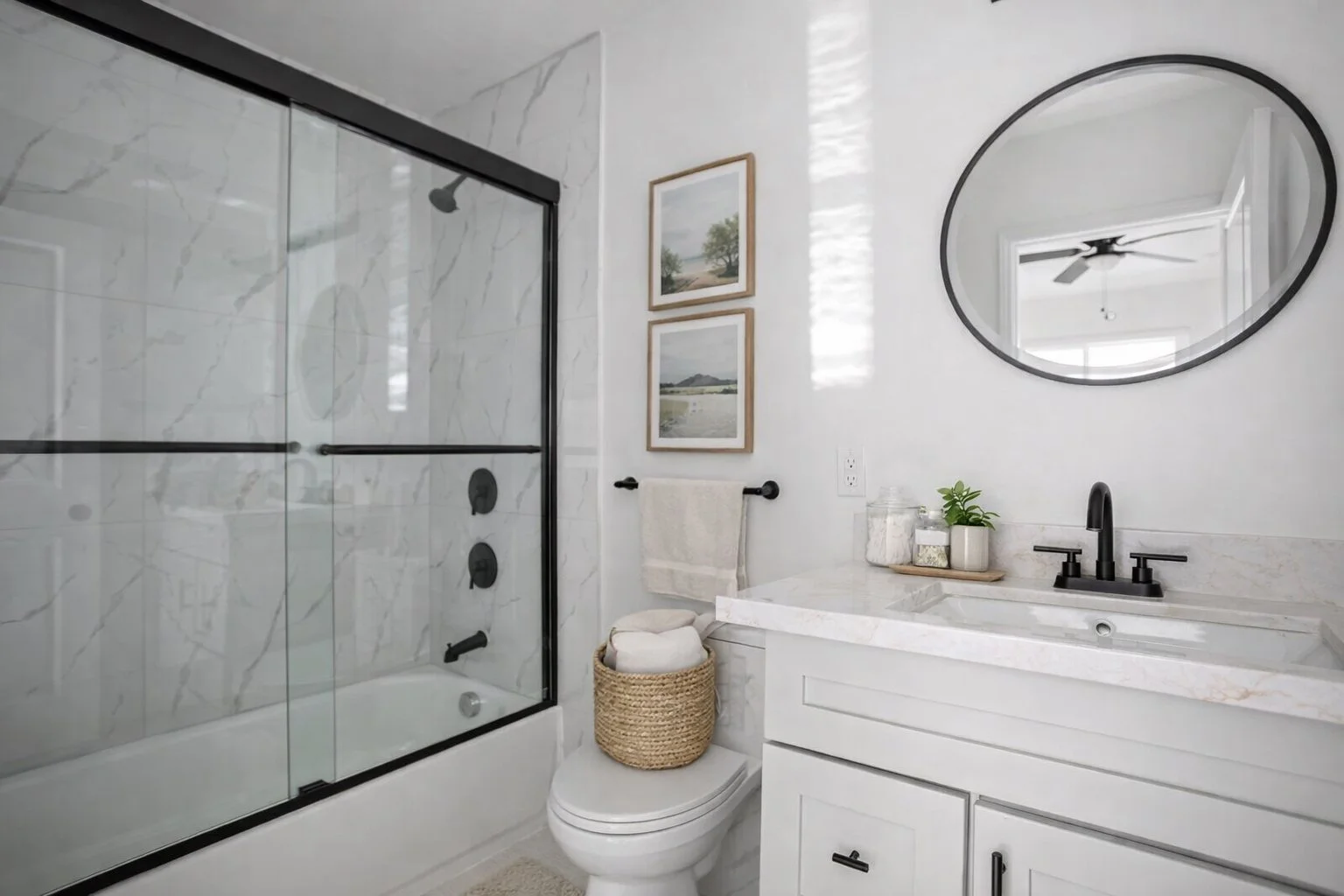 Bright modern bathroom with white marble accents, a glass shower with black frame, a white vanity with a black faucet, round mirror, framed pictures, and decorative items on the countertop.