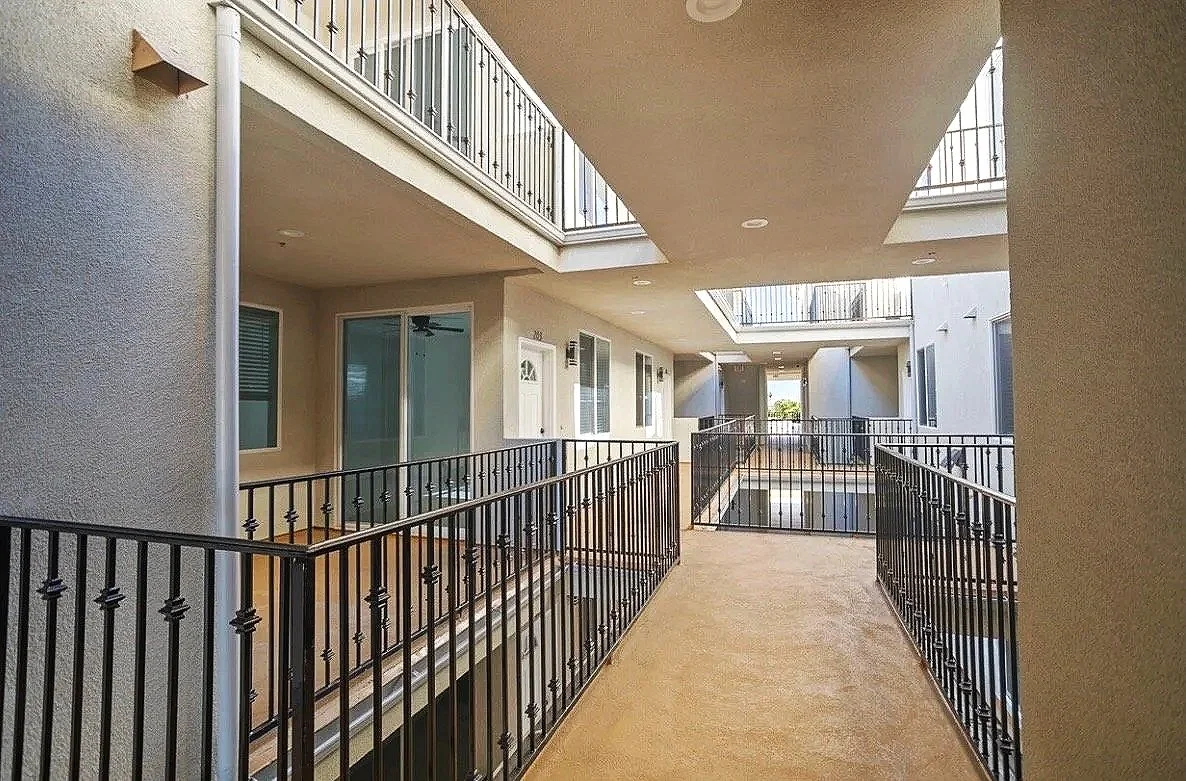 View of an apartment building corridor with beige walls, black metal railings, sliding glass doors, and balconies on upper floors, under a ceiling with ceiling lights.