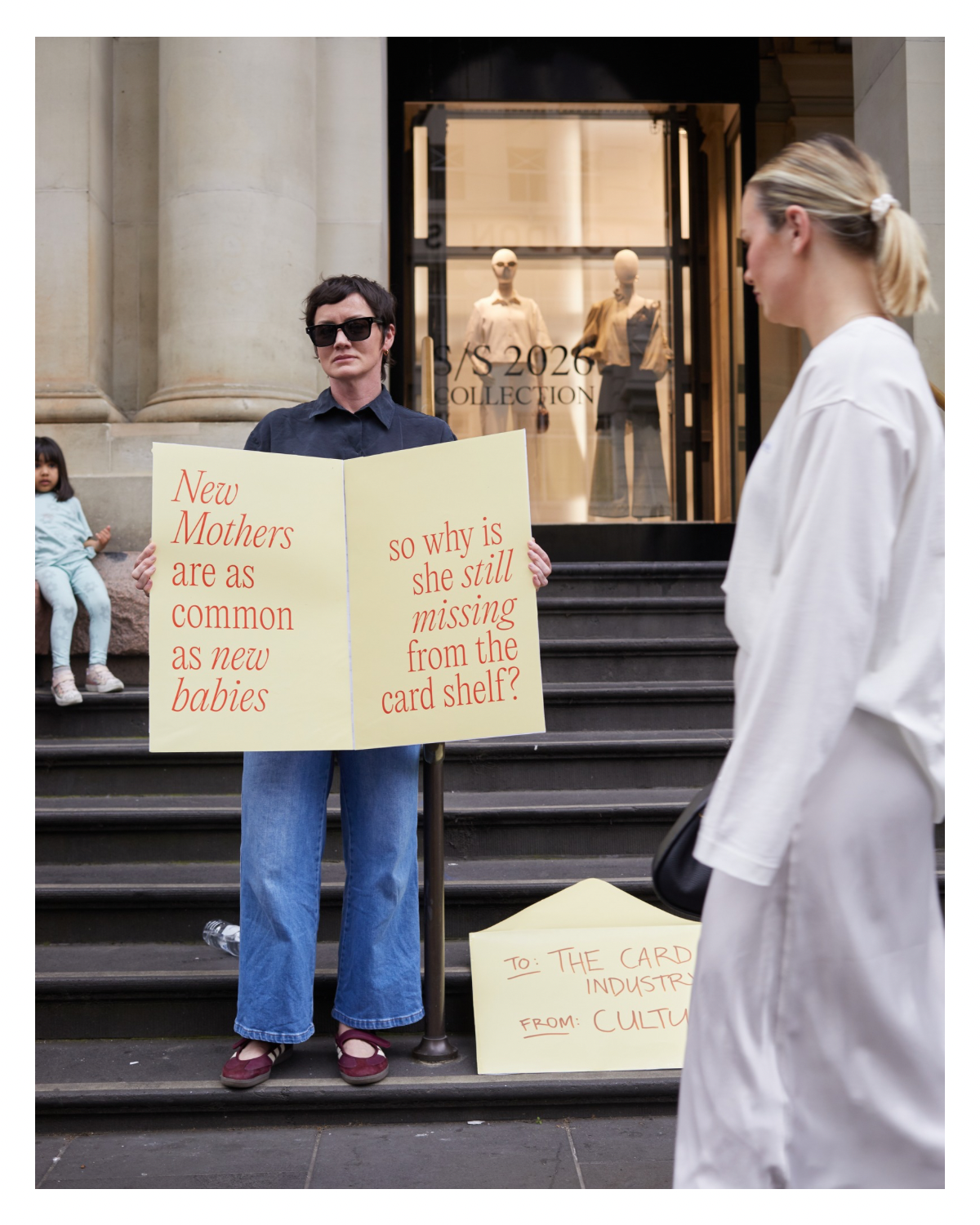 A woman with short dark hair, wearing sunglasses, a dark shirt, and blue jeans, holds a yellow sign with red text that reads, "New Mothers are as common as new babies so why is she still missing from the card shelf?" She is standing on steps outside a building with large columns. Another woman with blonde hair, wearing a white coat, stands in front of her. In the background, there are mannequins in a store window and children sitting on a bench.