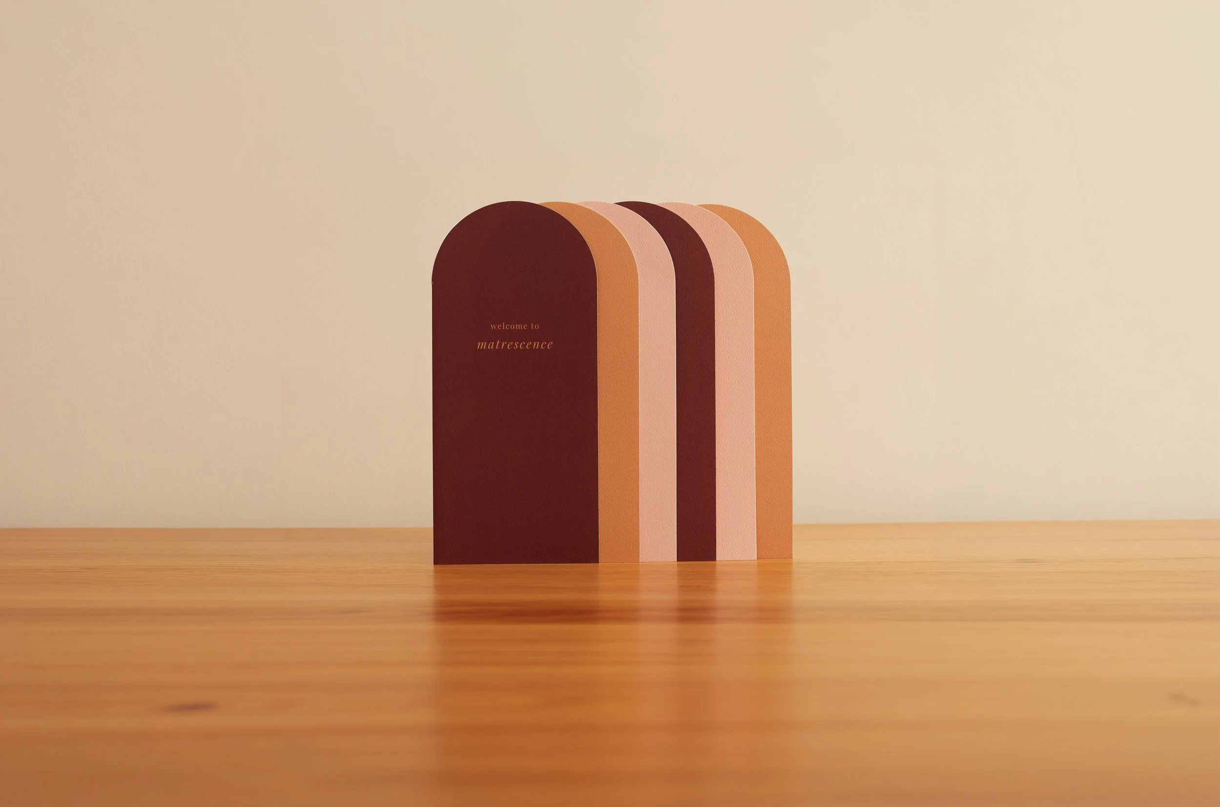 Six greeting cards with semicircular top edges in different shades of brown, beige, and peach, standing upright on a wooden surface against a beige background. The front card has the text 'welcome to matrescence' written in small, elegant font.