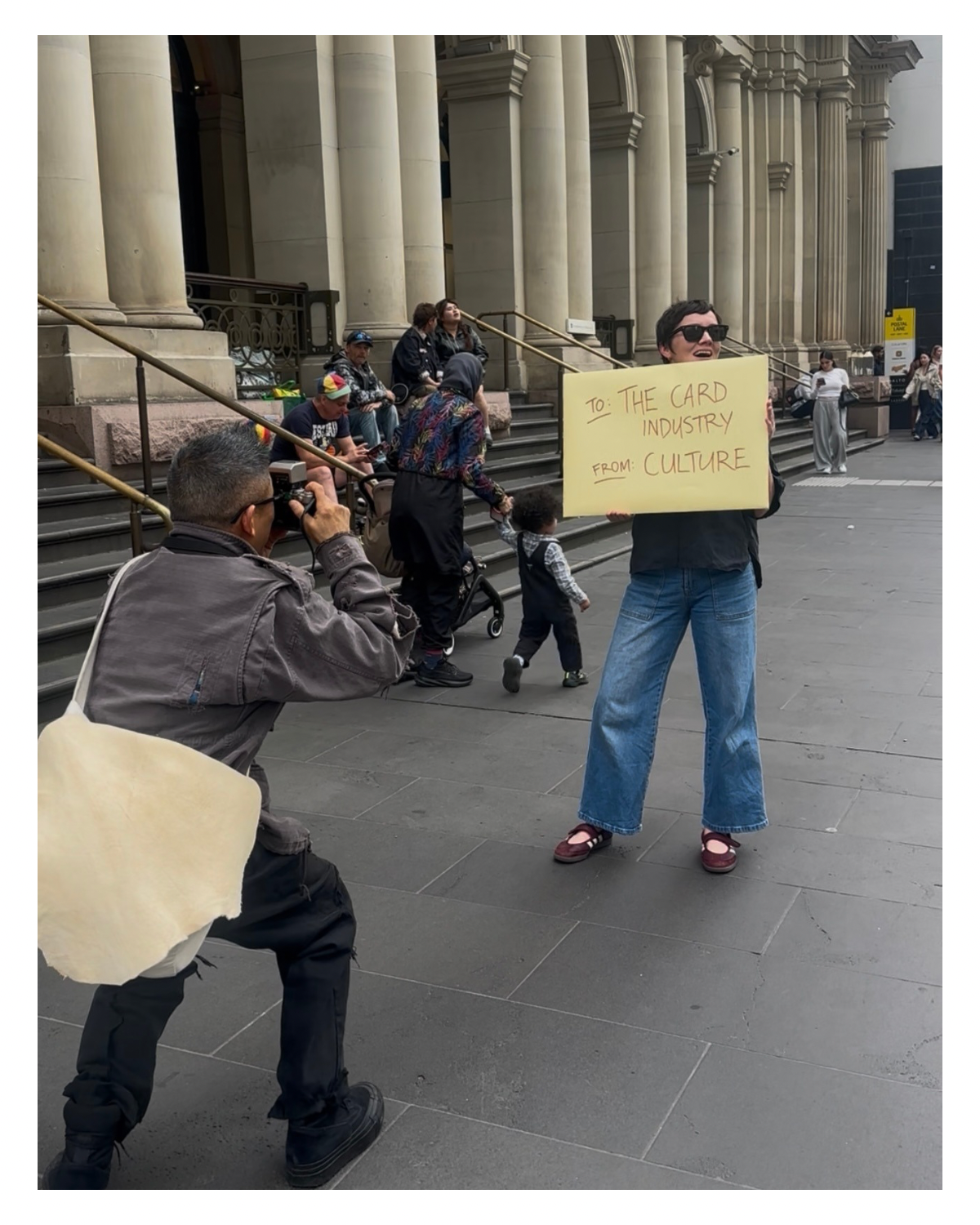 A man holding a large yellow sign that reads "To: The Card Industry, From: Cultures". He is wearing sunglasses, a dark shirt, and jeans. A photographer is taking his picture while various people are sitting and walking in the background on a city street with large building columns.