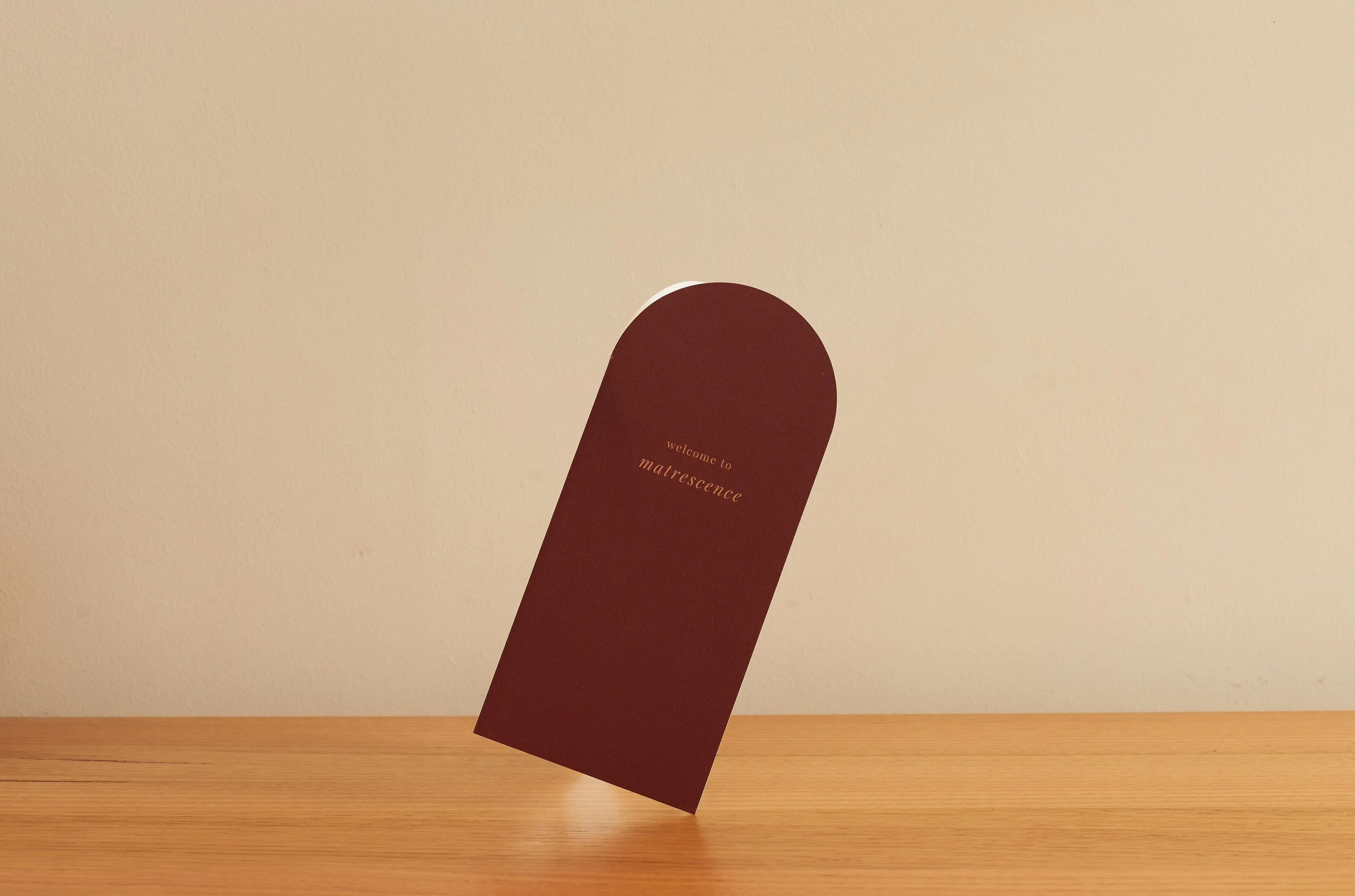 A maroon invitation card with gold text that reads 'welcome to matrescence' is displayed on a wooden table against a plain beige wall.