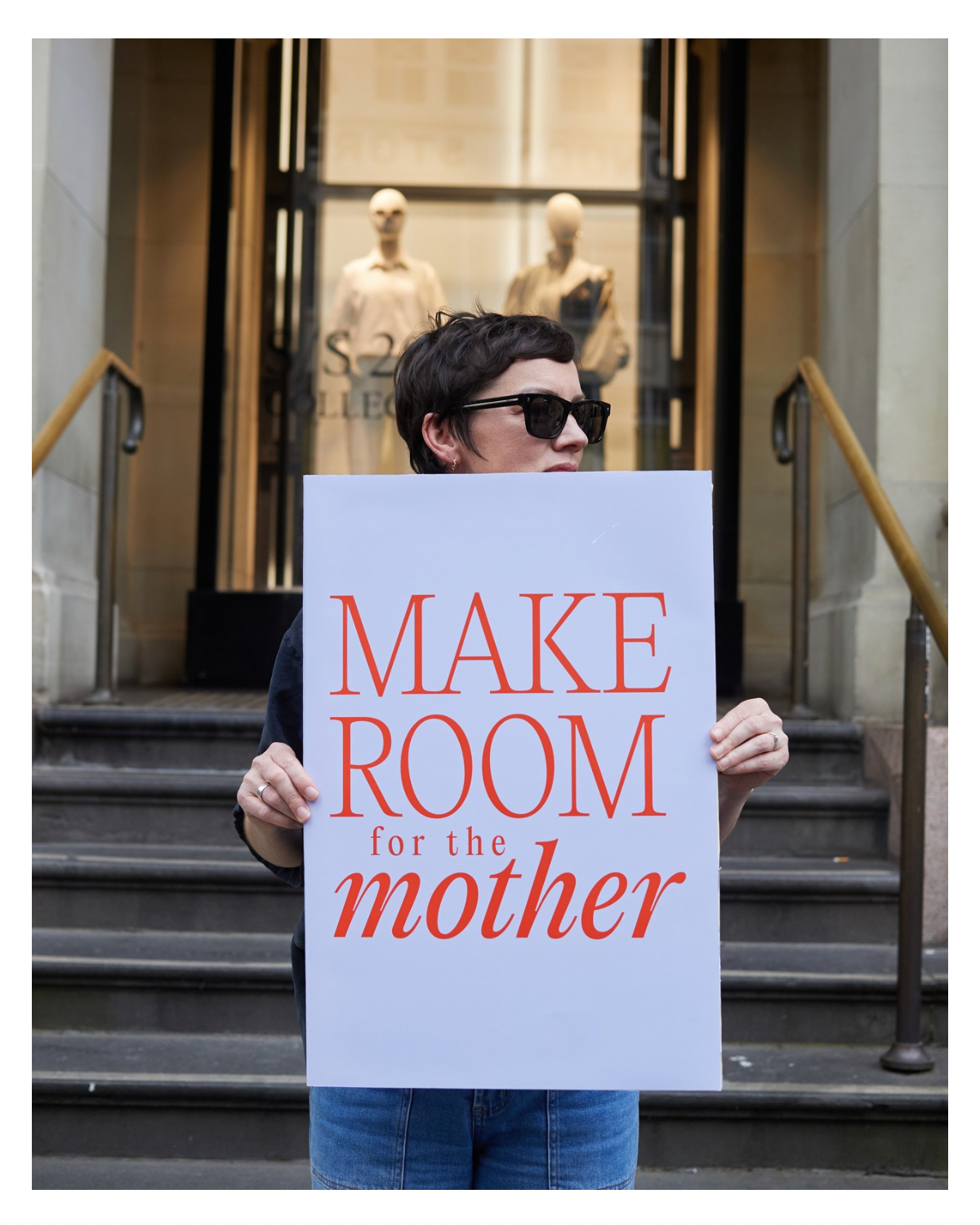Person holding a sign that reads 'MAKE ROOM for the mother' standing on city stairs in front of a store window display with mannequins.