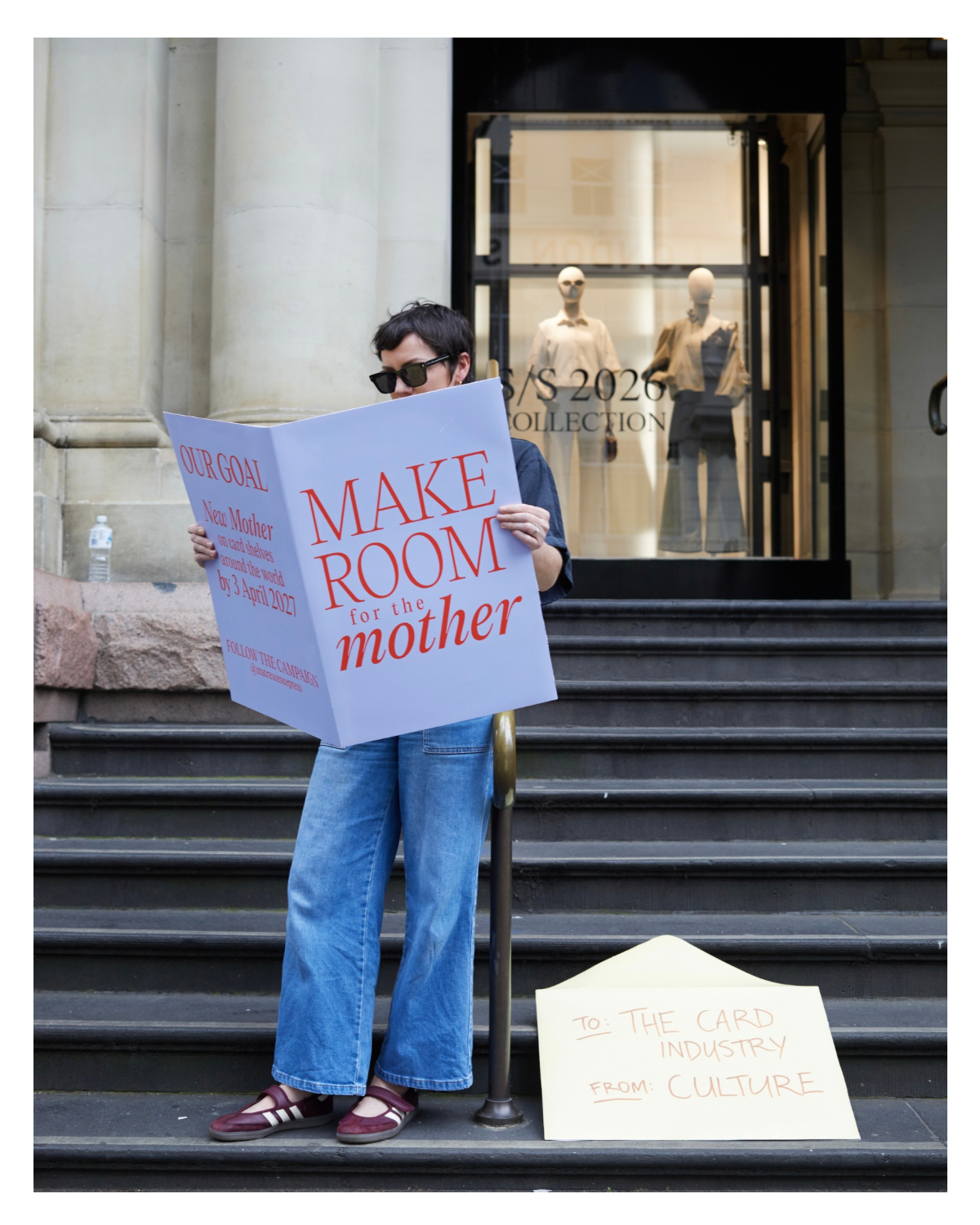 A person standing on steps holding a large sign that says, 'MAKE ROOM for the mother,' with additional text about a goal to create for new mothers. and fashion collection signage. There is also aon the steps that says, 'TO: THE CARD INDUSTRY FROM: CULTURE.'