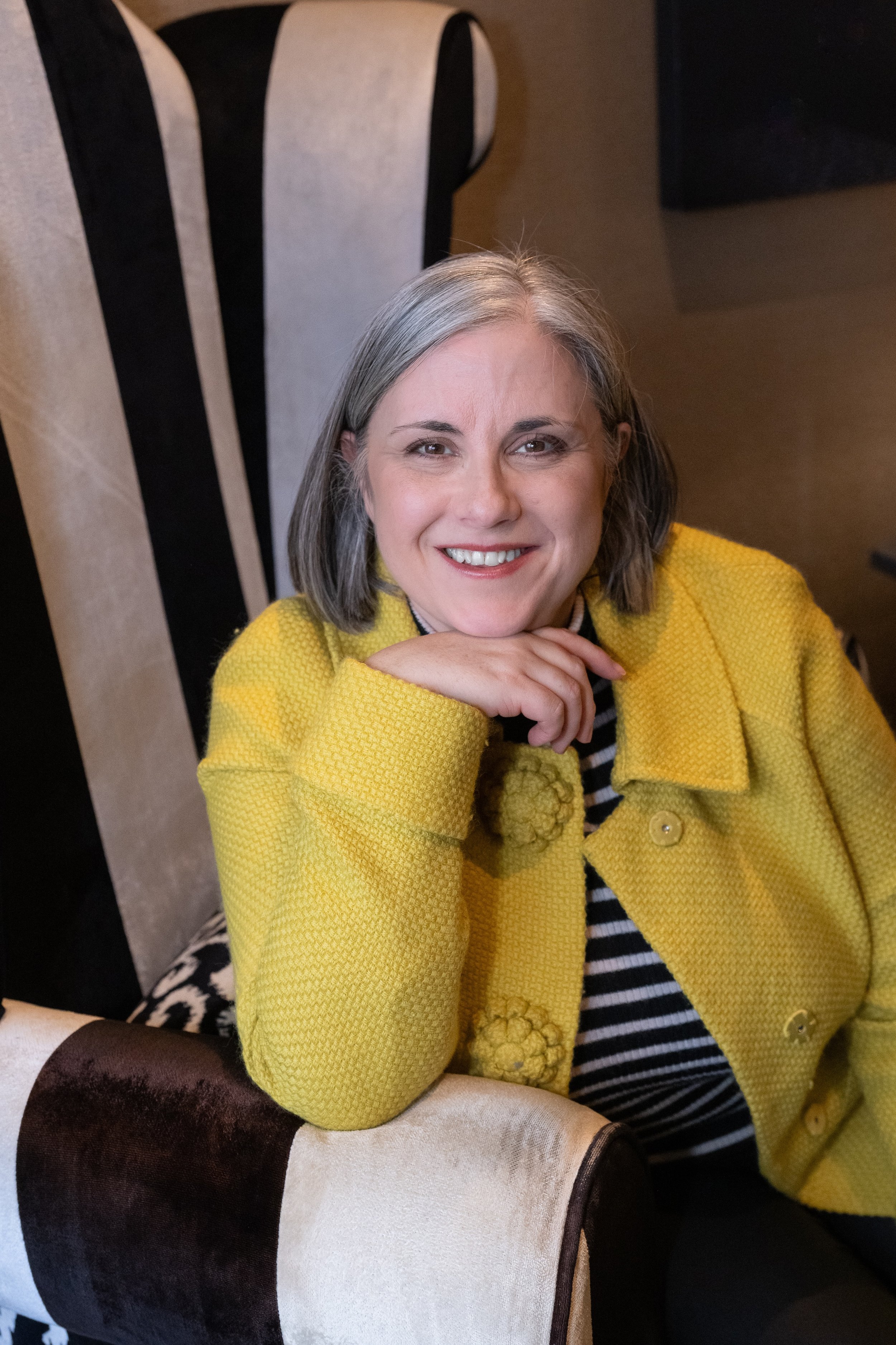 Coach Jo - A woman with grey hair, smiling and wearing a yellow textured jacket, sitting on a patterned upholstered chair.