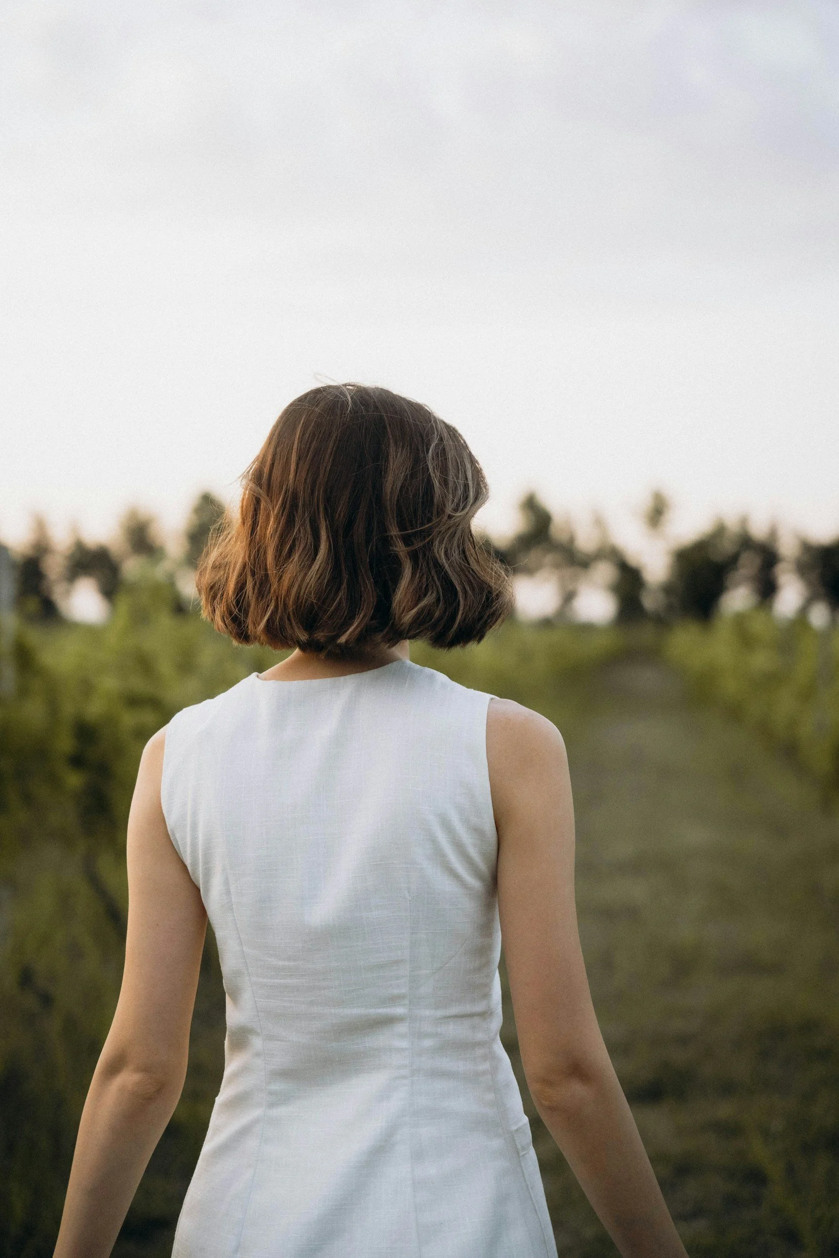 Rear view of a woman with short brown hair wearing a sleeveless white dress standing outdoors in a green landscape with trees in the background.